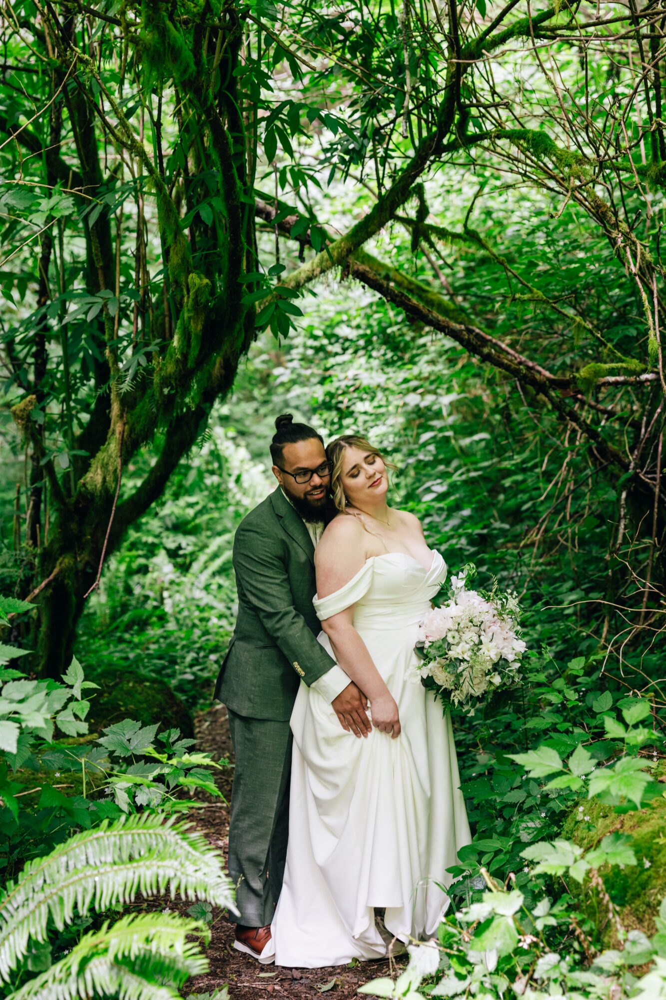 A couple in wedding attire standing in a lush green forest, holding a bouquet of flowers, with dense foliage around them.
