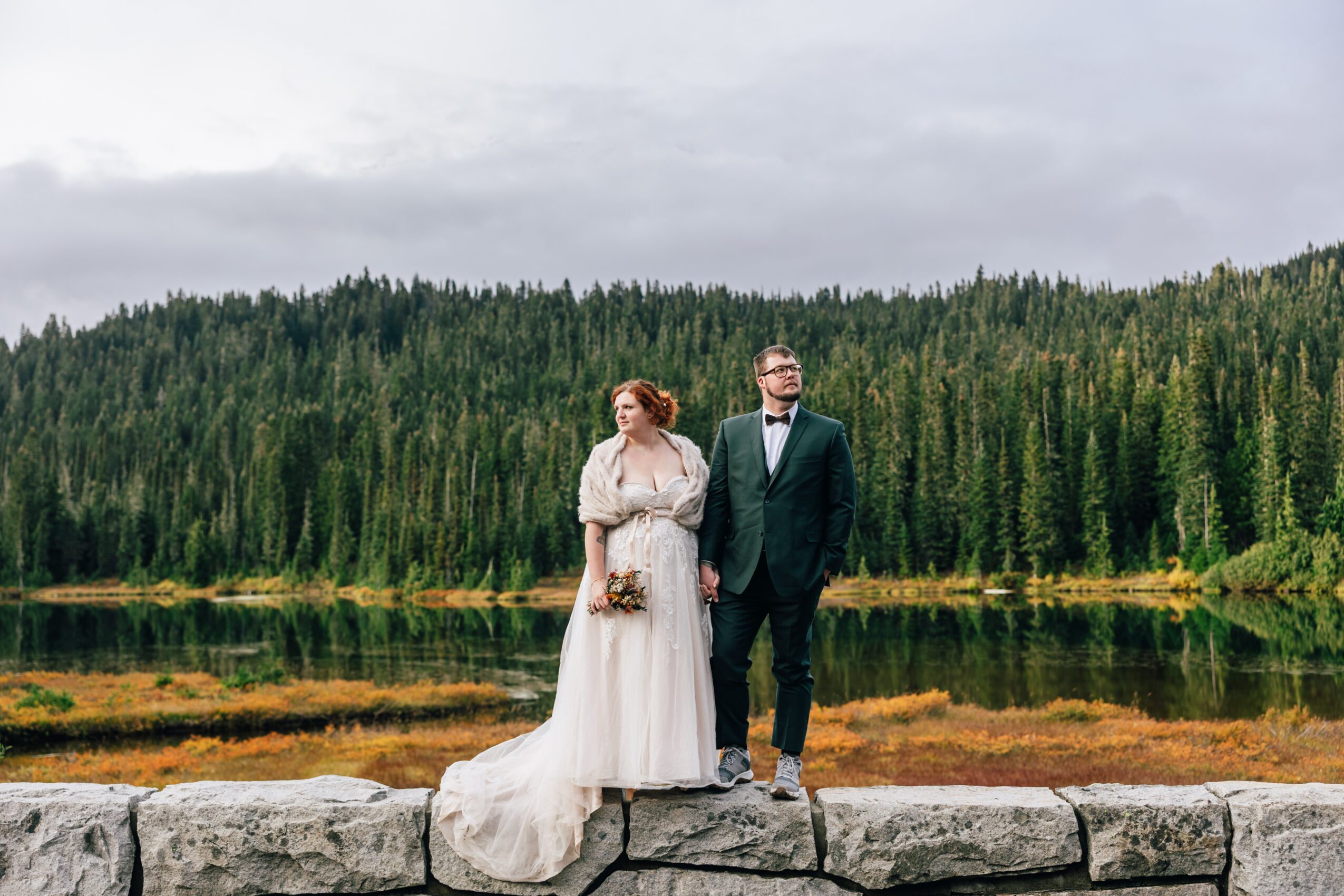 A bride and groom stand on a stone ledge by a lake with a forested background, dressed in wedding attire.