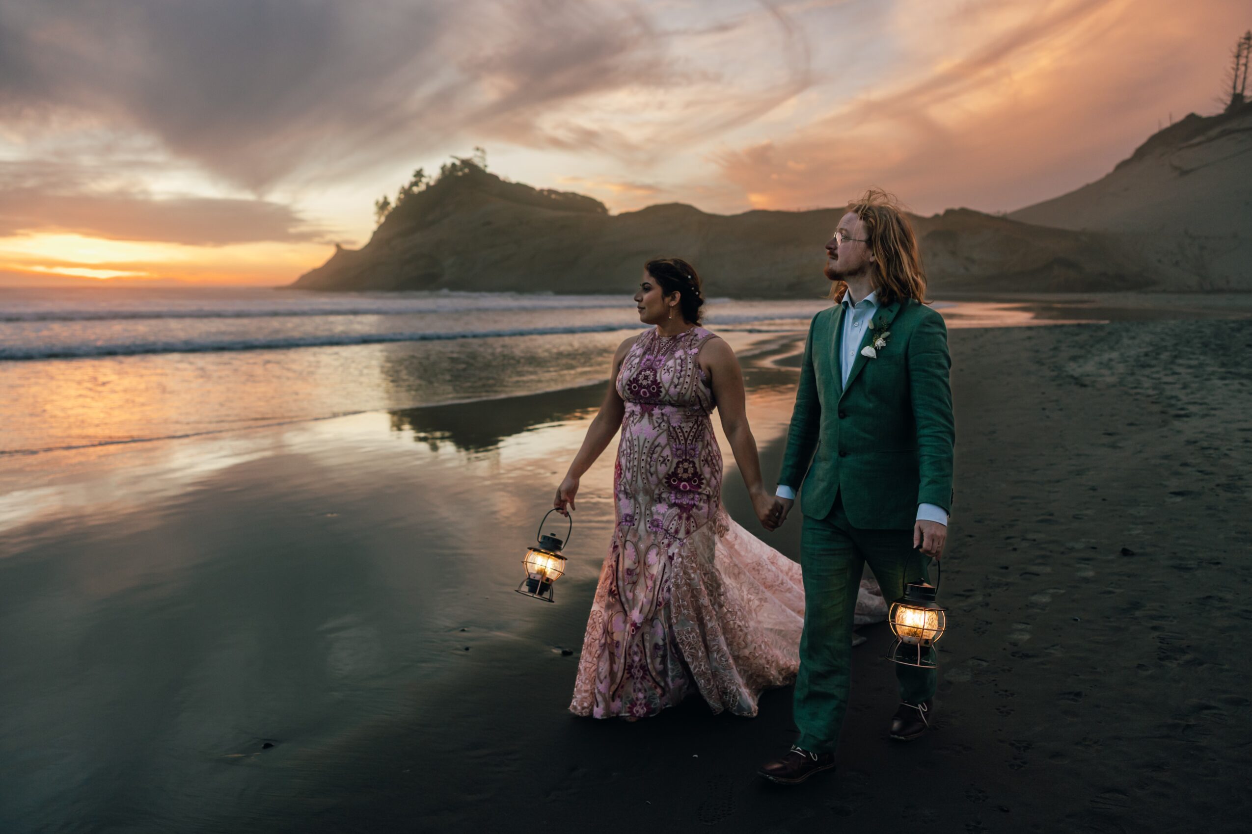 A couple in formal attire walking on a beach at sunset, holding lanterns, with a hilly landscape in the background.