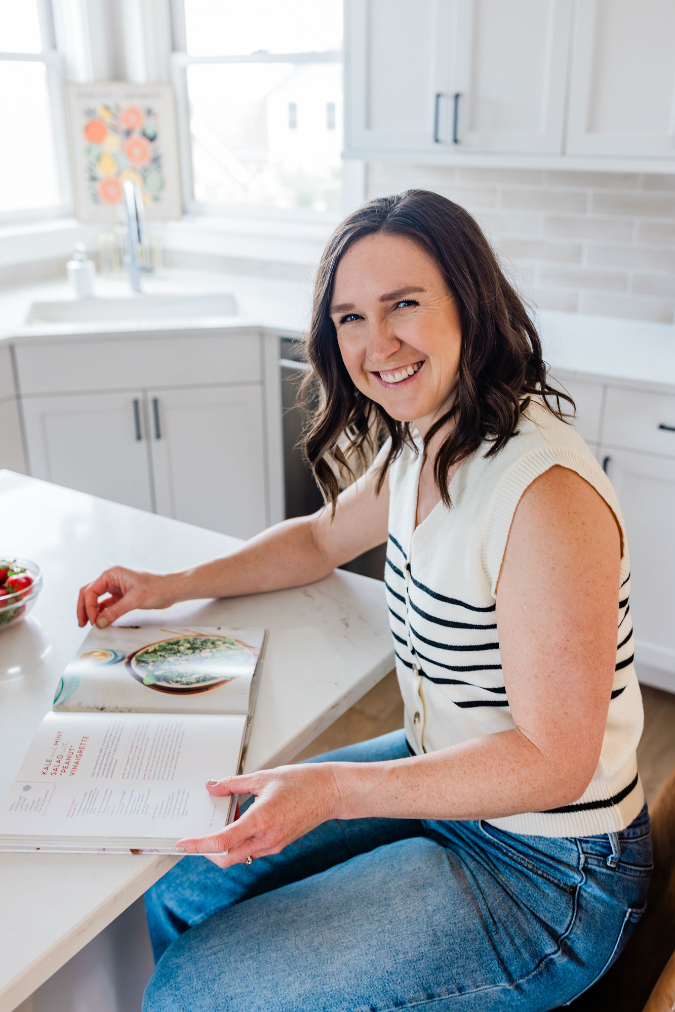 Woman with dark hair smiling, sitting at kitchen counter with open book, in bright modern kitchen.