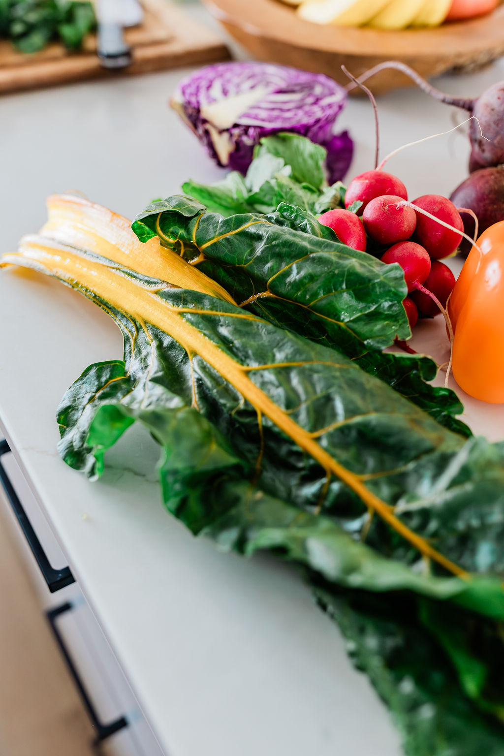 Fresh leafy greens, radishes, and colorful vegetables on a kitchen counter, with a blurred background of a cutting board and purple cabbage.