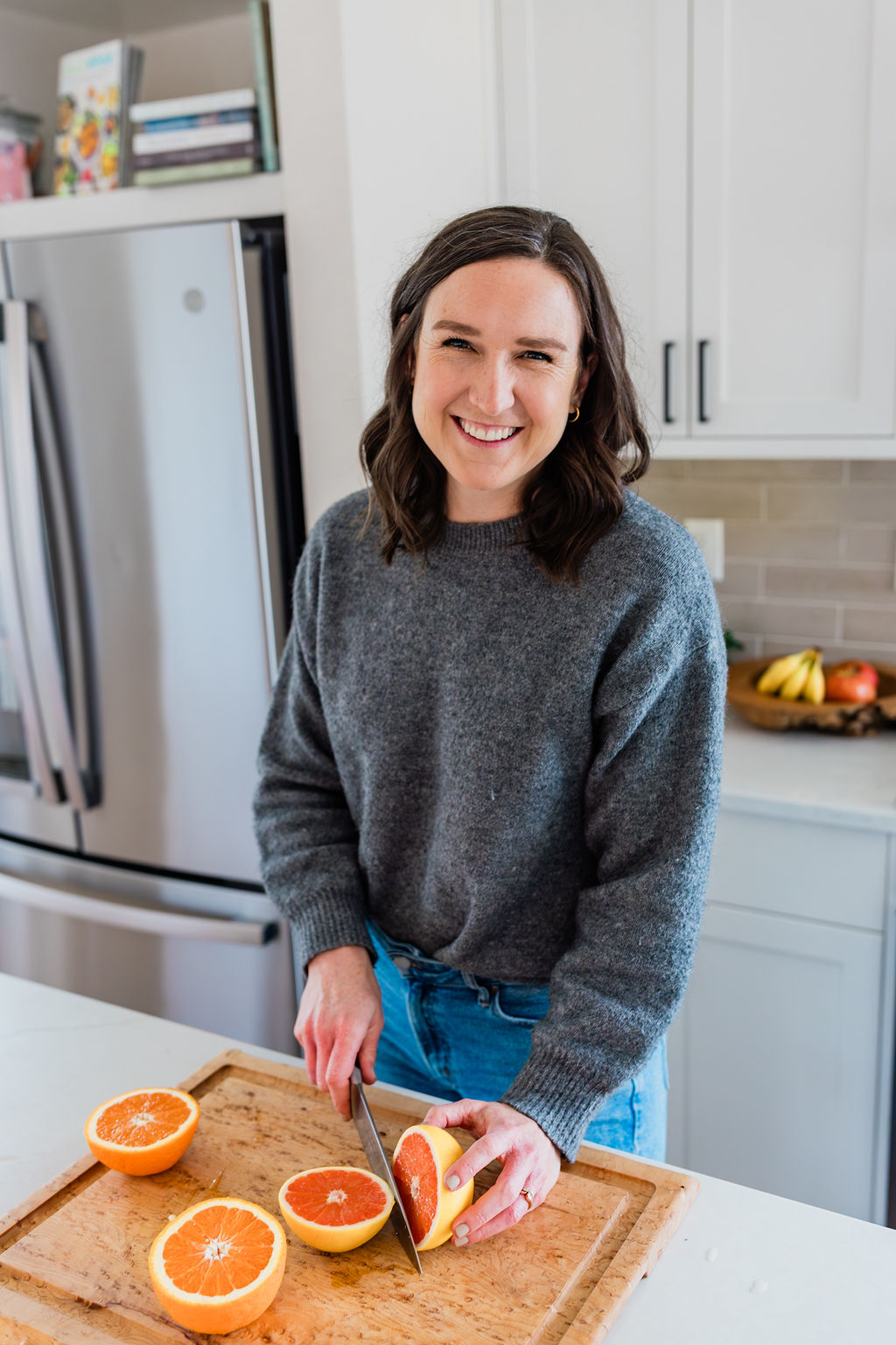 Woman smiling in a kitchen, cutting a grapefruit on a wooden cutting board, with oranges and a knife nearby.