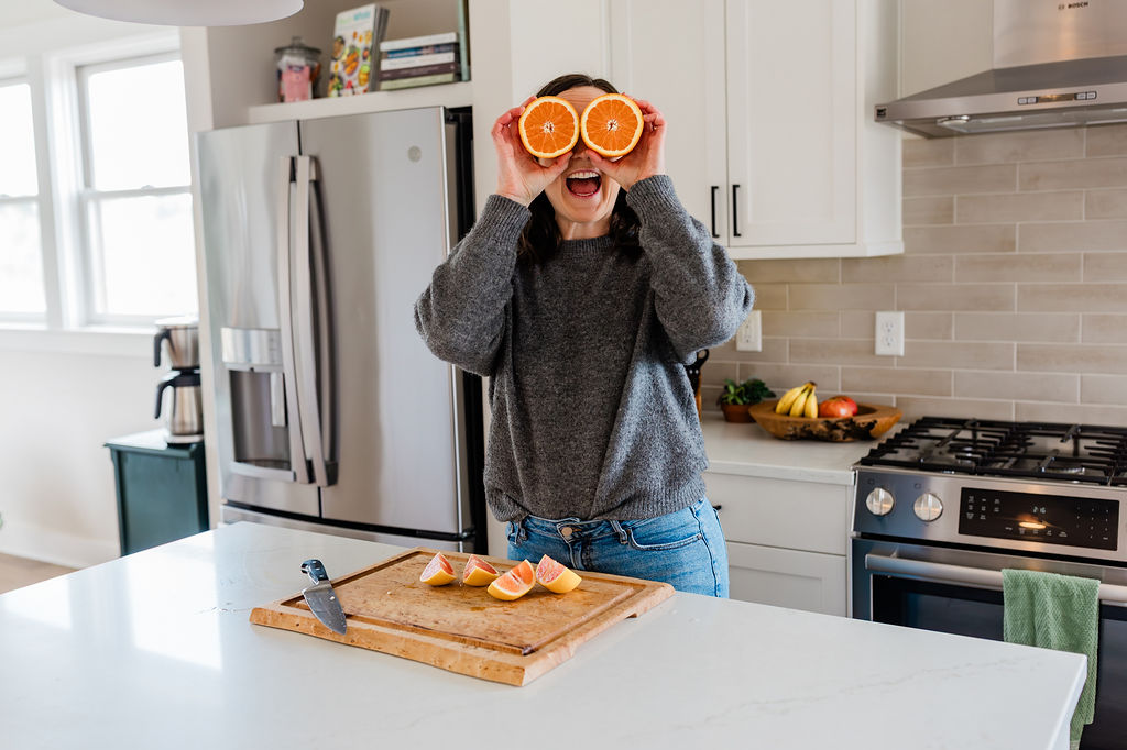 Person in a kitchen holding orange slices over eyes, smiling, with a cutting board and fruit on the counter.