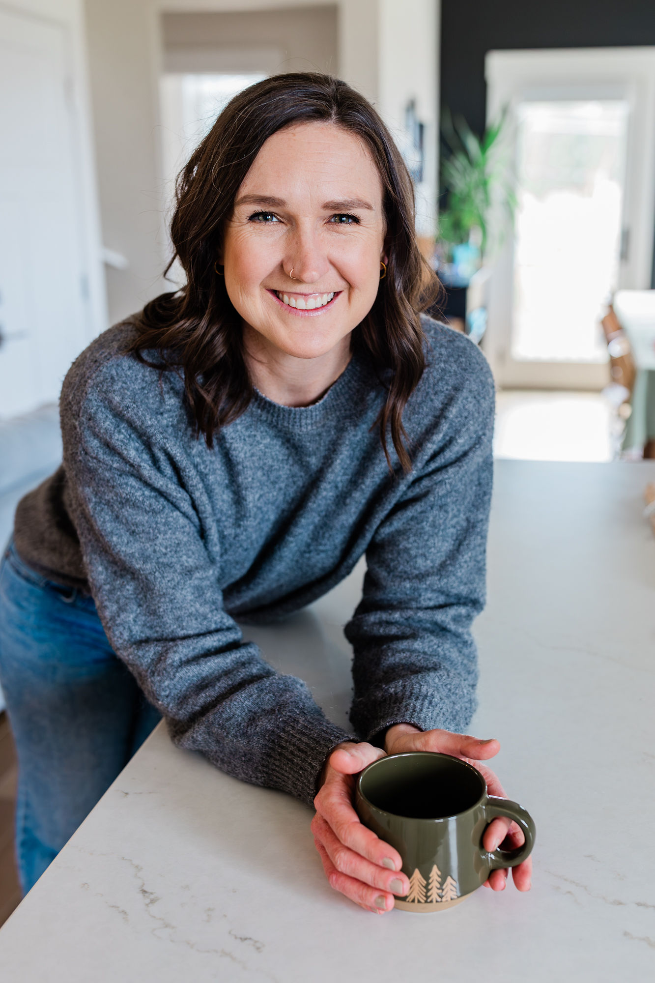 Woman with shoulder-length dark hair smiling, holding a mug, in a bright kitchen, wearing a blue sweater.