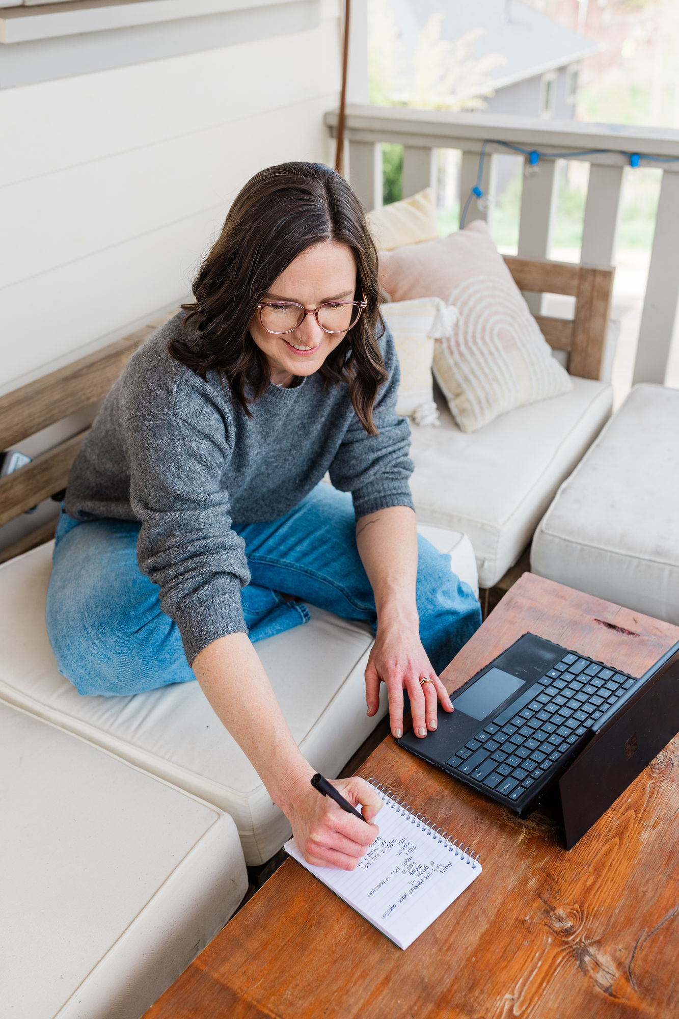 Woman with glasses sitting on a couch, writing on a notepad, with a laptop nearby, in a bright room.