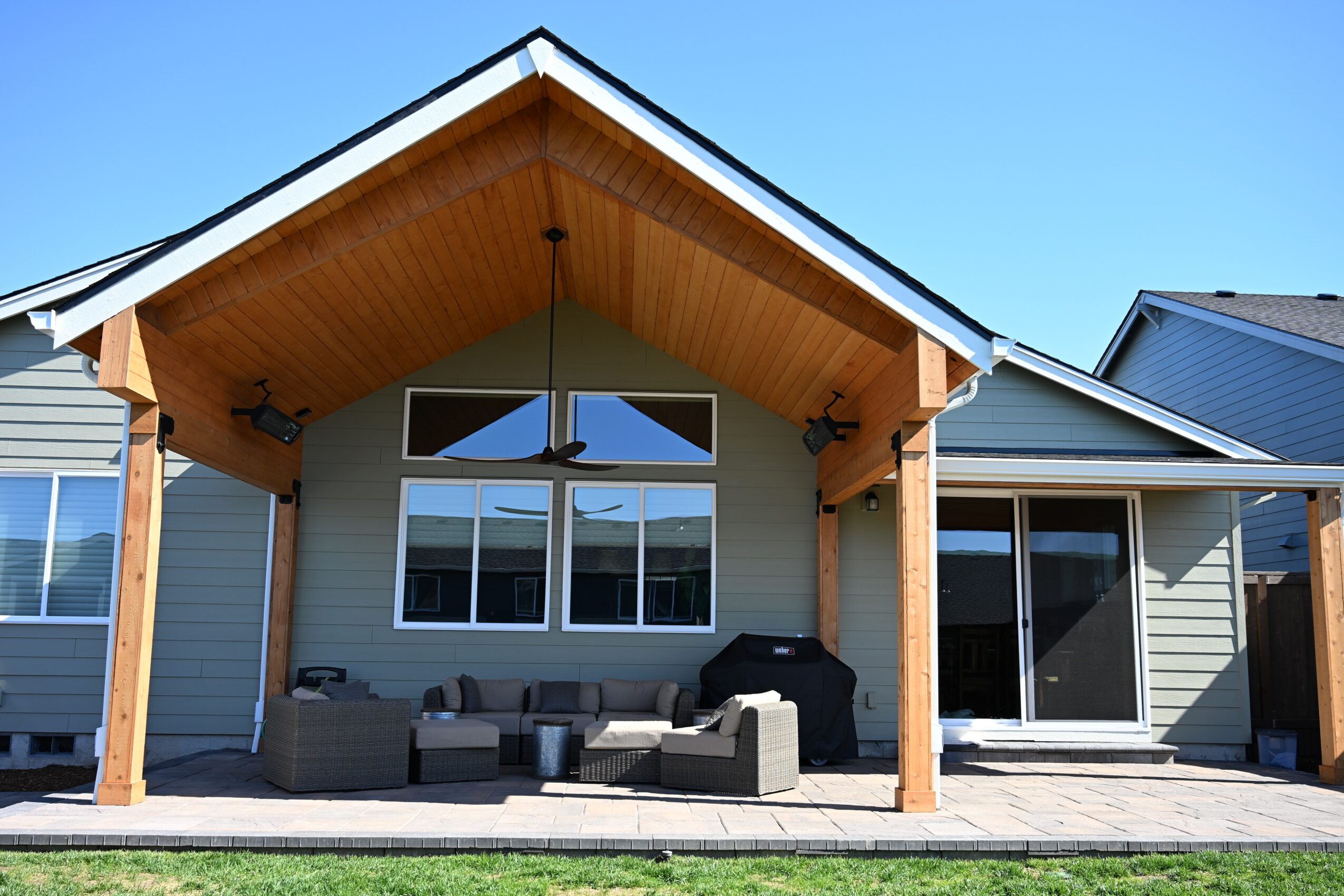 Front view of a house with a large covered porch, wooden beams, and outdoor seating, under a clear blue sky.