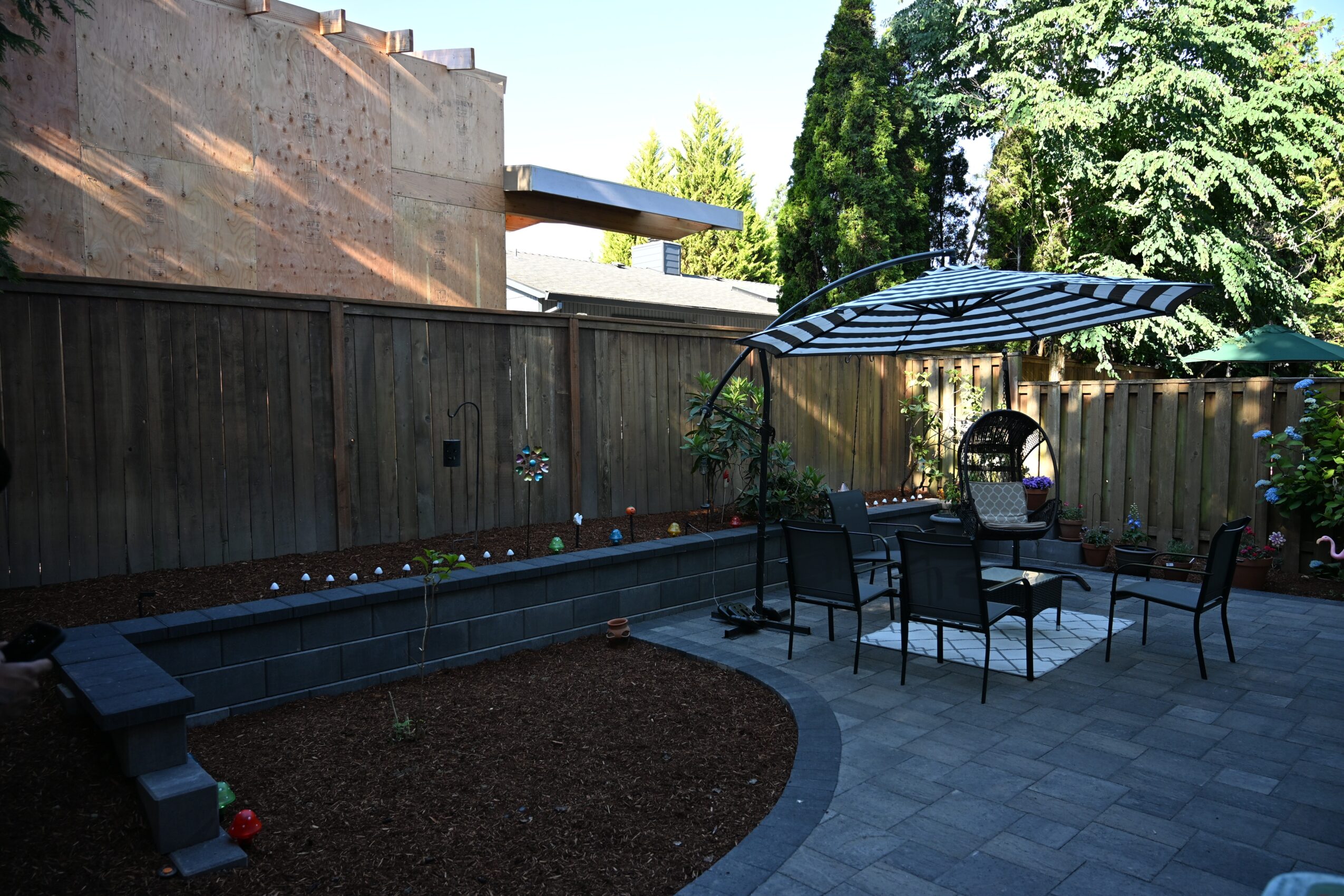 Backyard patio with a table, chairs, umbrella, and a small pool, enclosed by a wooden fence with trees in the background.