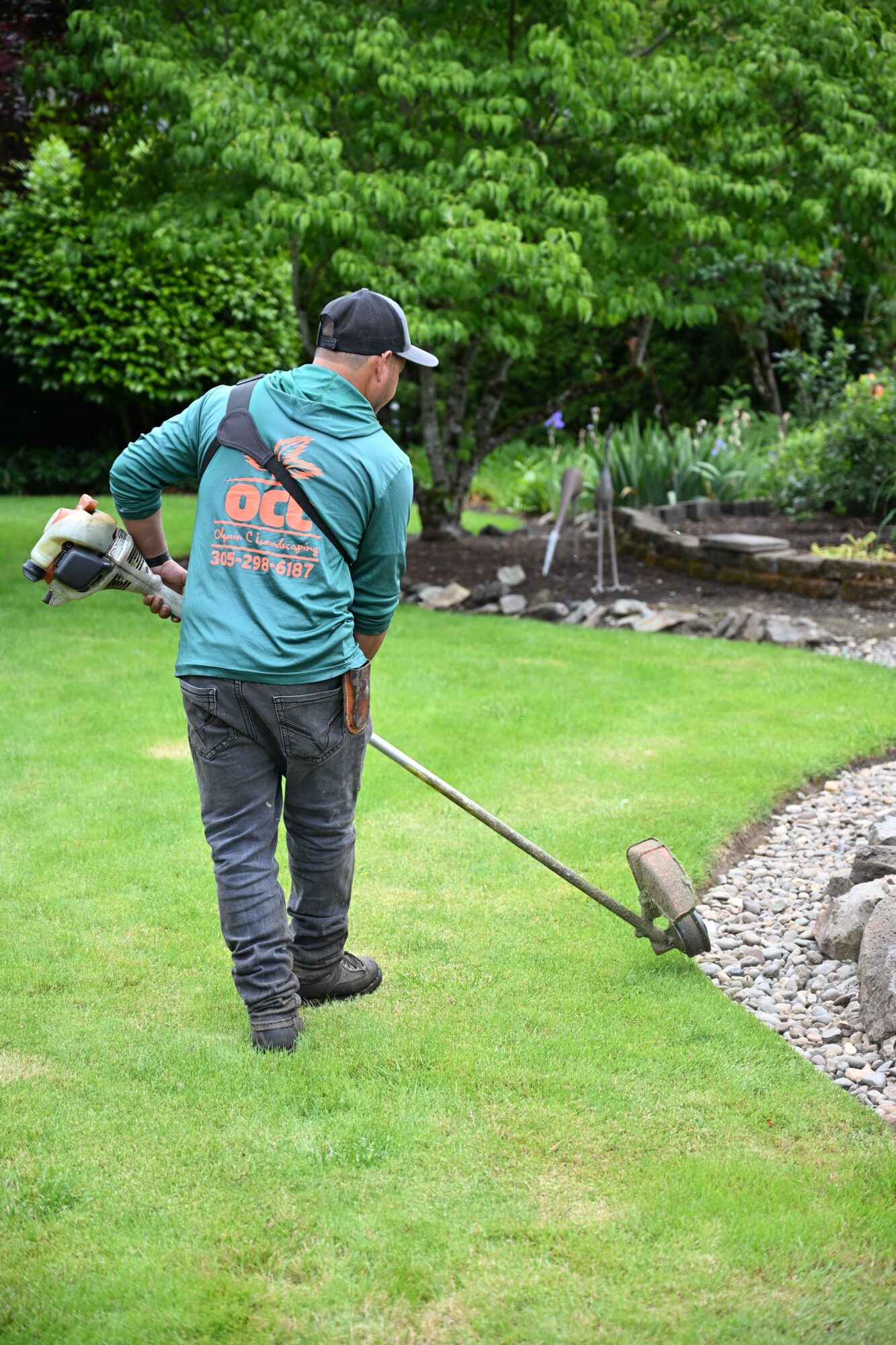 Person raking gravel in a garden with green grass and trees, wearing a blue hoodie and black cap.