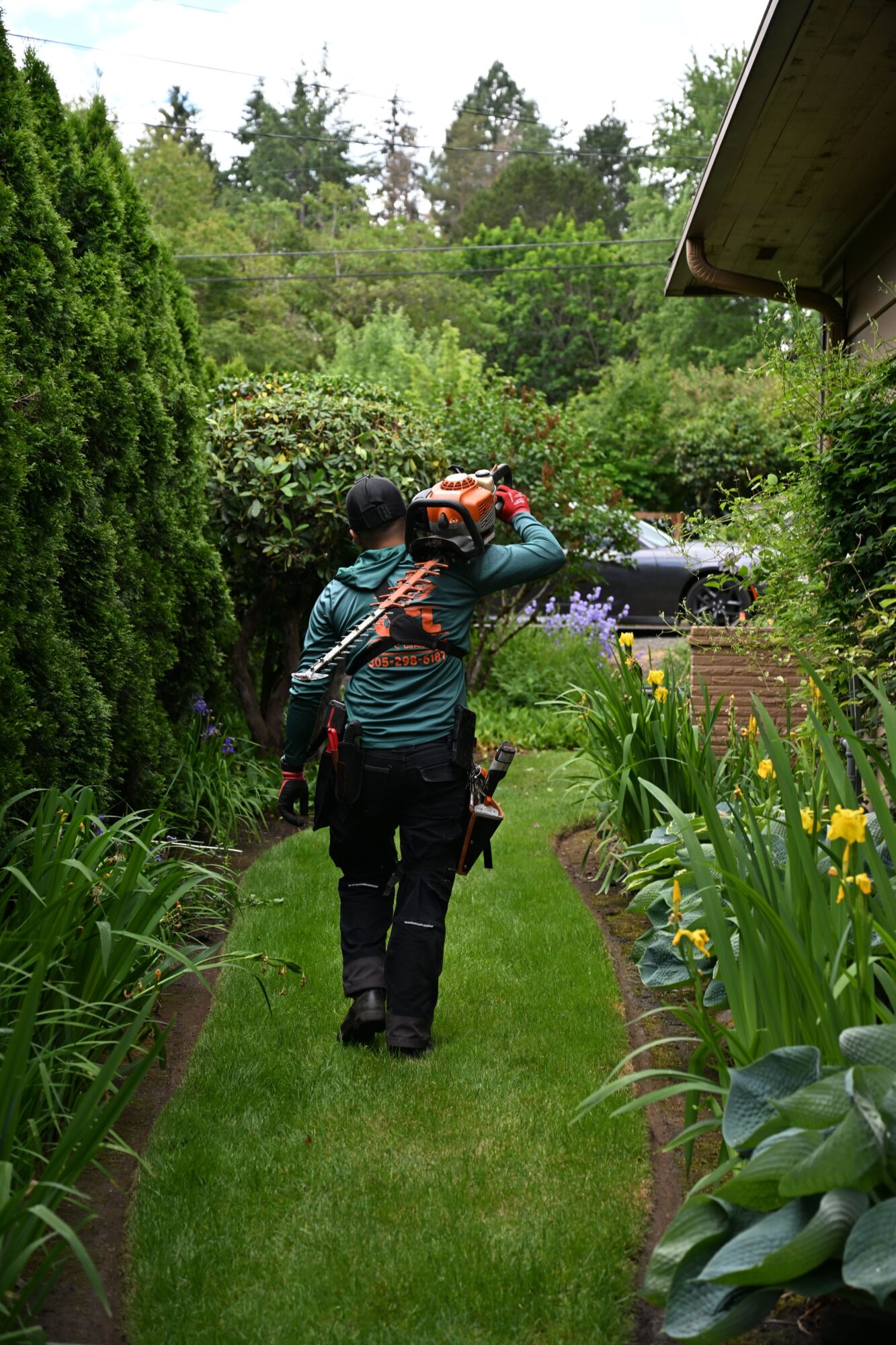 Person with backpack walking on a garden path surrounded by green plants and flowers.