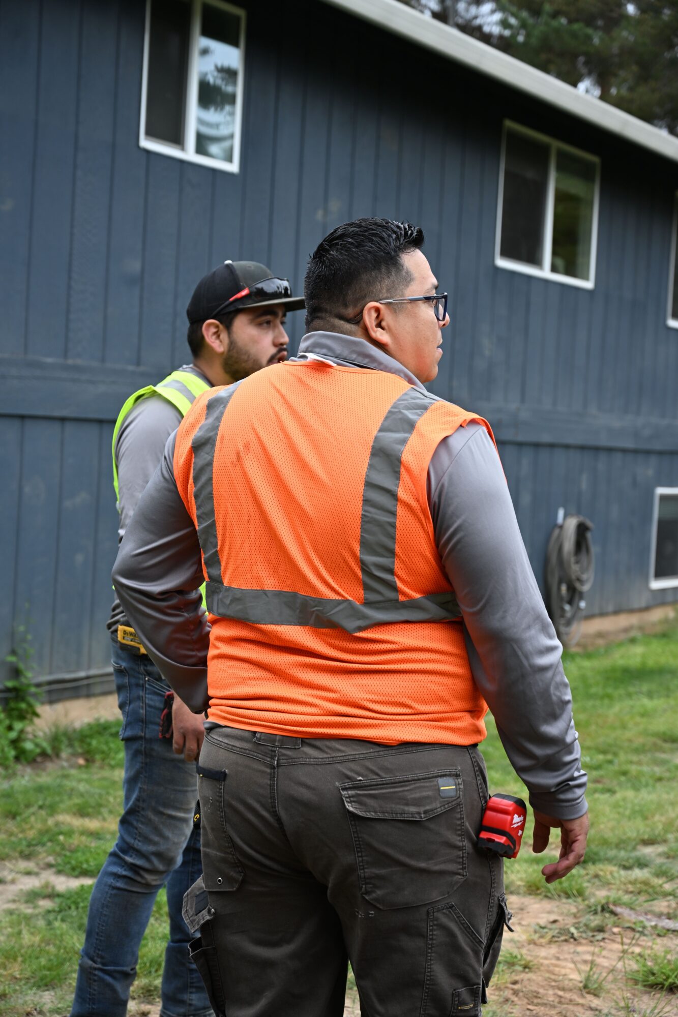Two men standing outdoors near a blue building, one wearing an orange safety vest and glasses, the other in a gray jacket and cap.