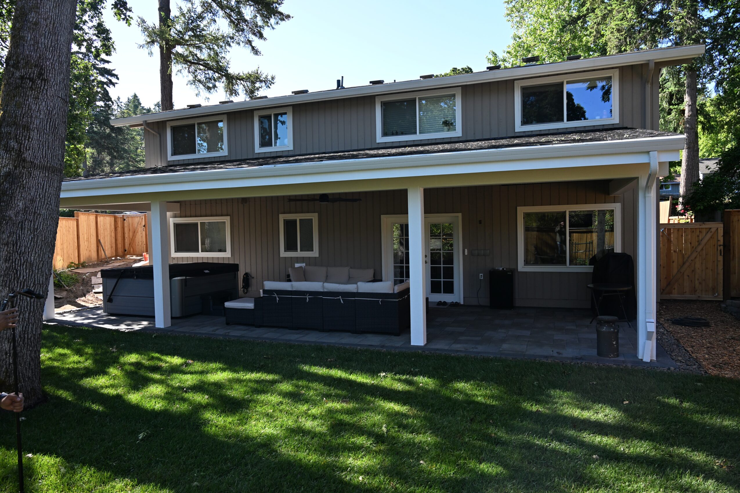 Two-story house with a covered porch, outdoor seating, and a grassy yard, surrounded by trees and a wooden fence.