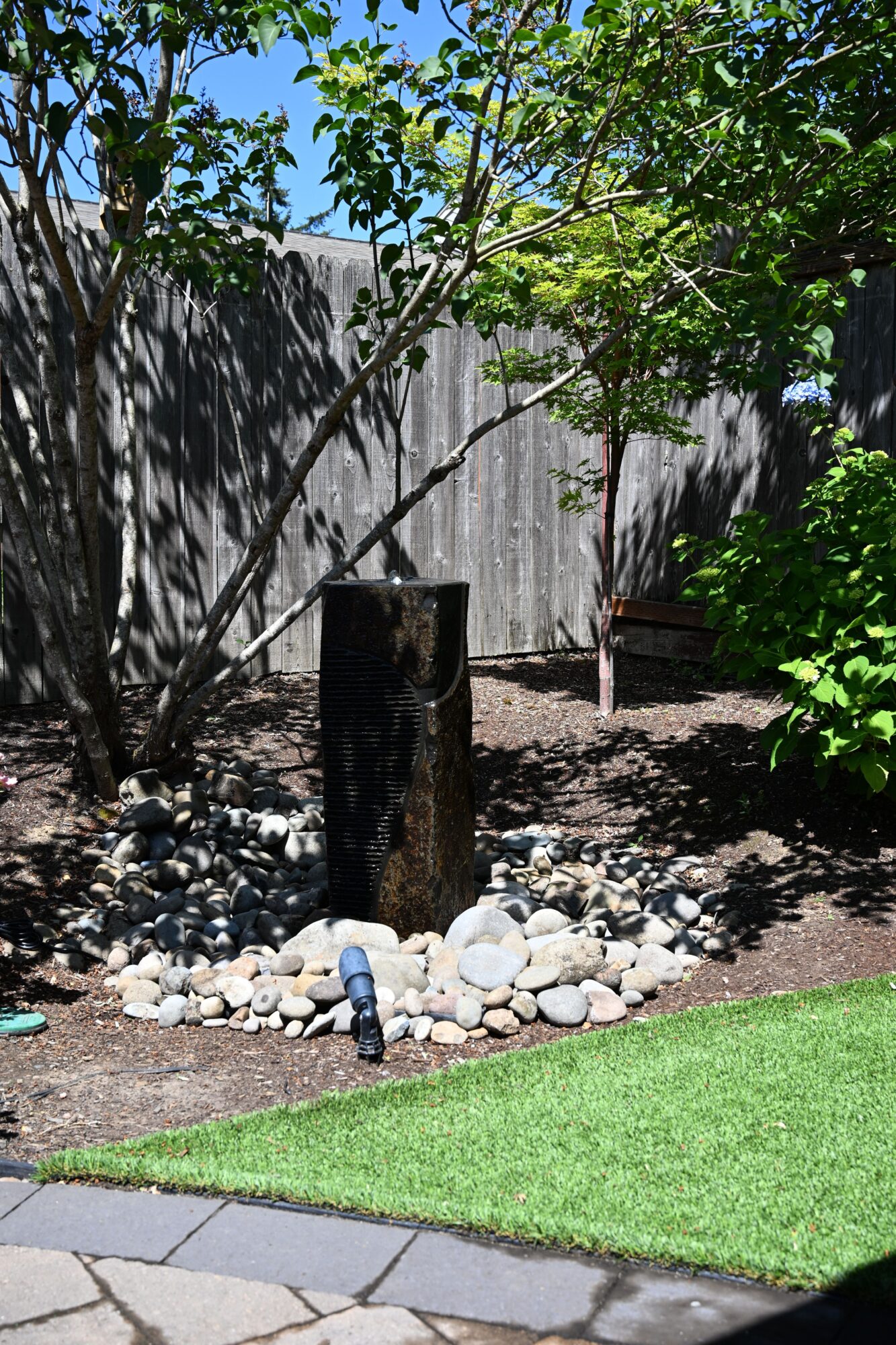 A backyard garden with a tree, rocks, a wooden fence, and a patch of artificial grass, under a blue sky.