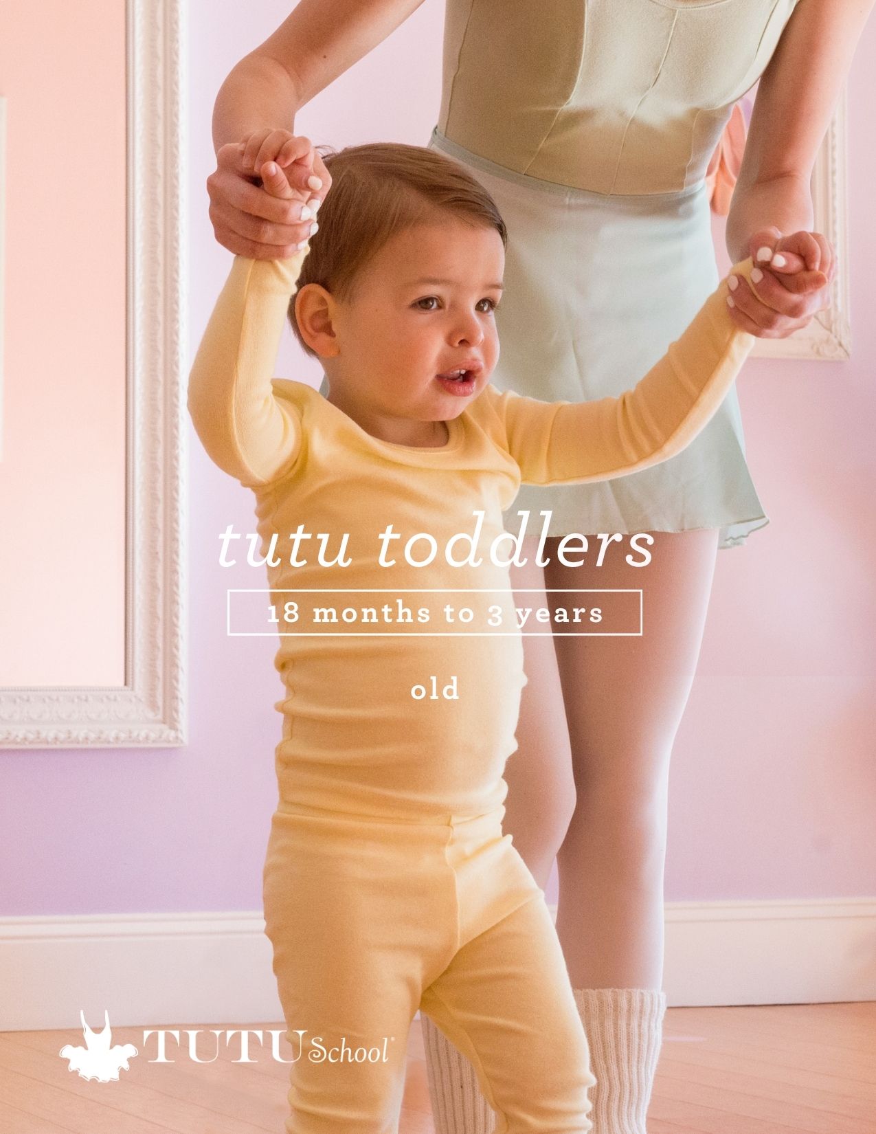 Young child in yellow outfit holding hands with an adult, standing indoors with pink walls and wooden floor.