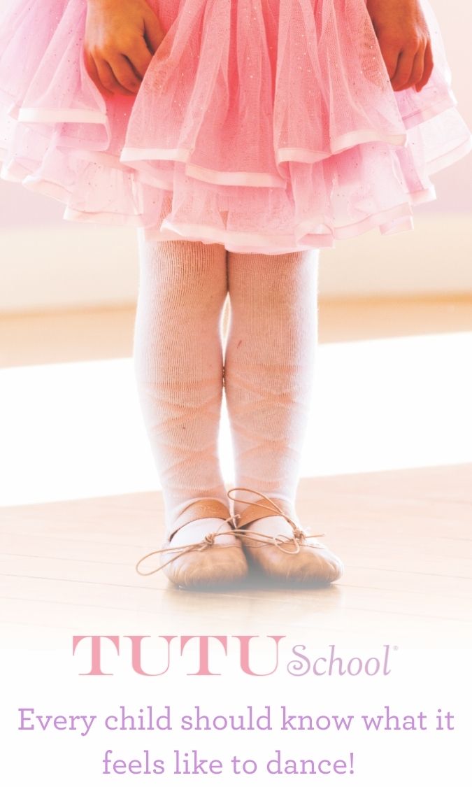Close-up of a child's legs in a pink tutu dress, white tights, and ballet shoes, standing on a wooden floor.