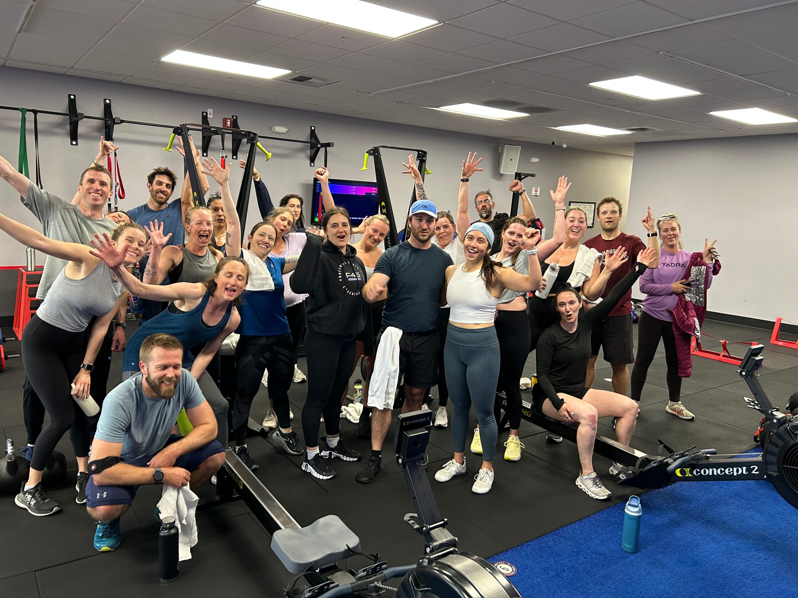 Group of people in a gym, some with arms raised, smiling, with exercise equipment around them.