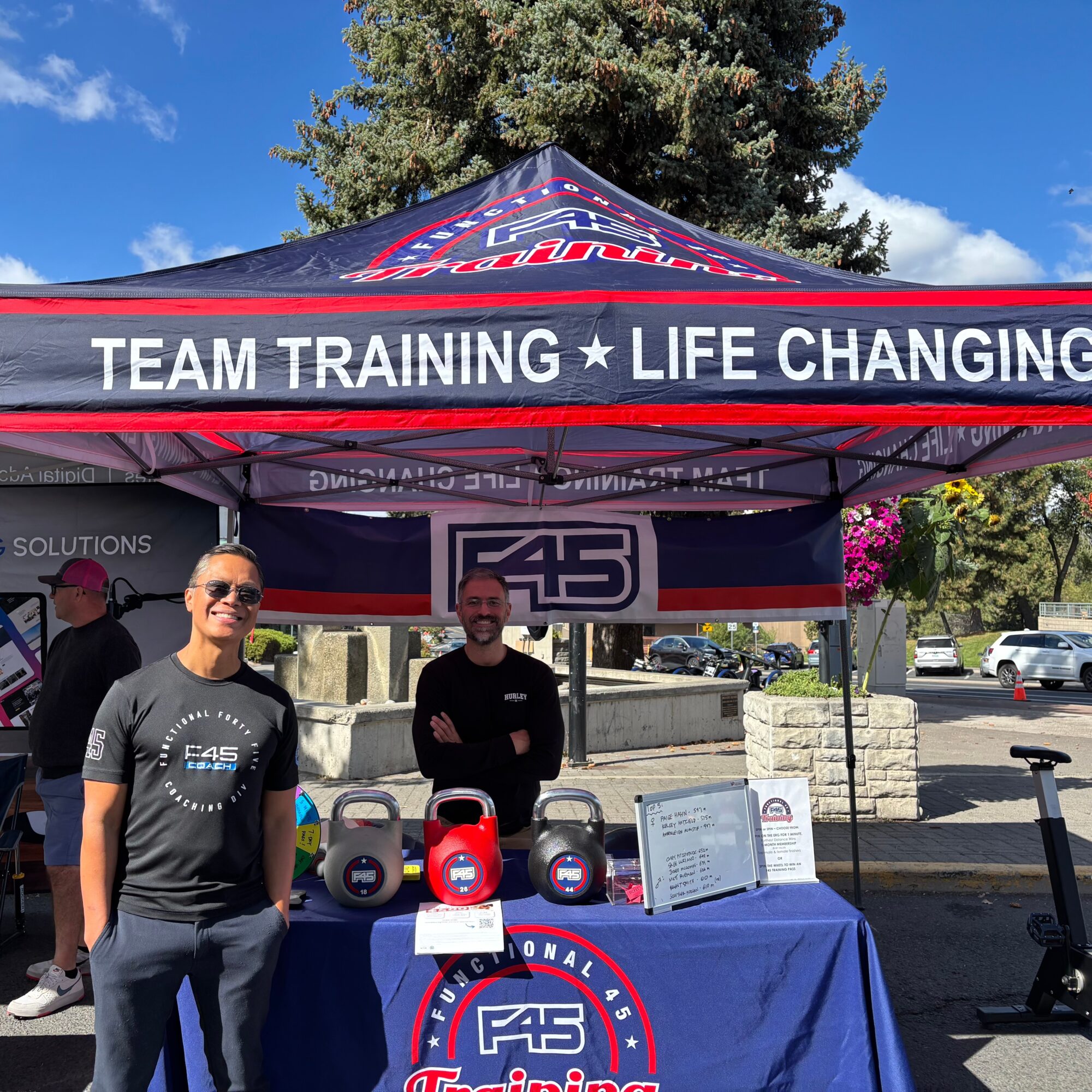 Two men standing behind a table under a canopy with the logo 'F45' and kettlebells, outdoors on a sunny day.