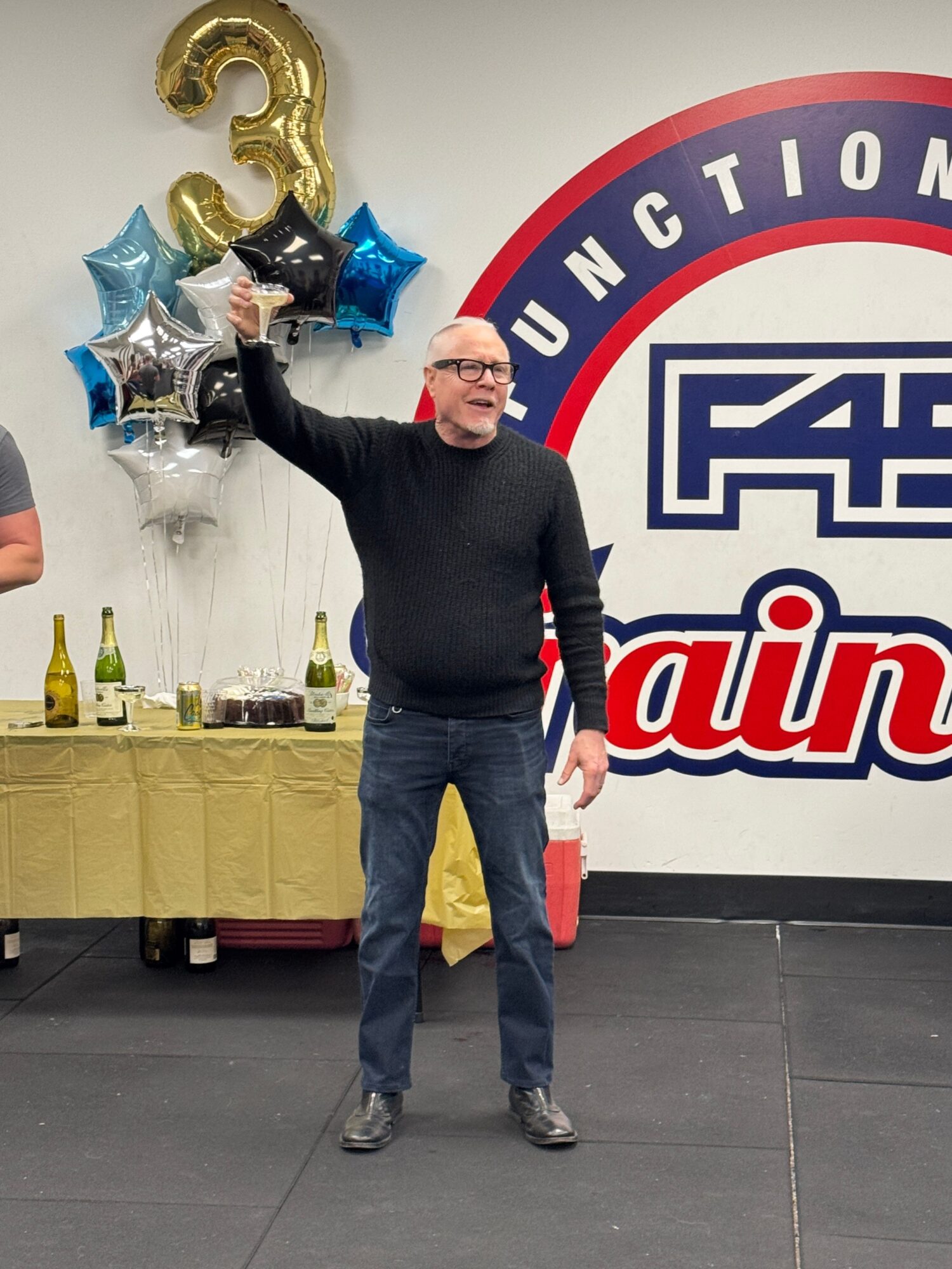 Person standing indoors near a table with balloons and drinks, holding a trophy, with a large sign in the background.