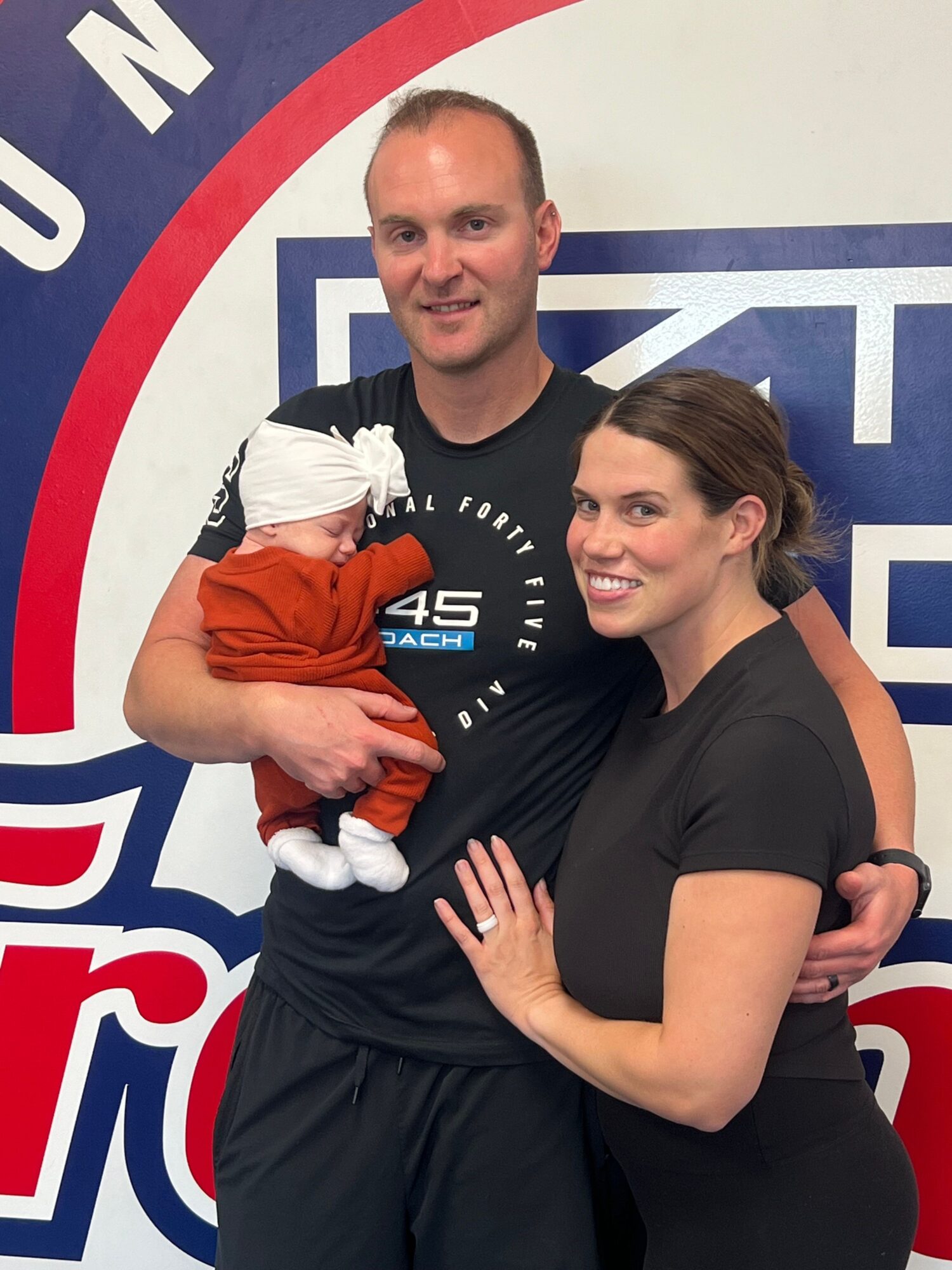 A smiling woman and a man holding a baby in front of a logo background.