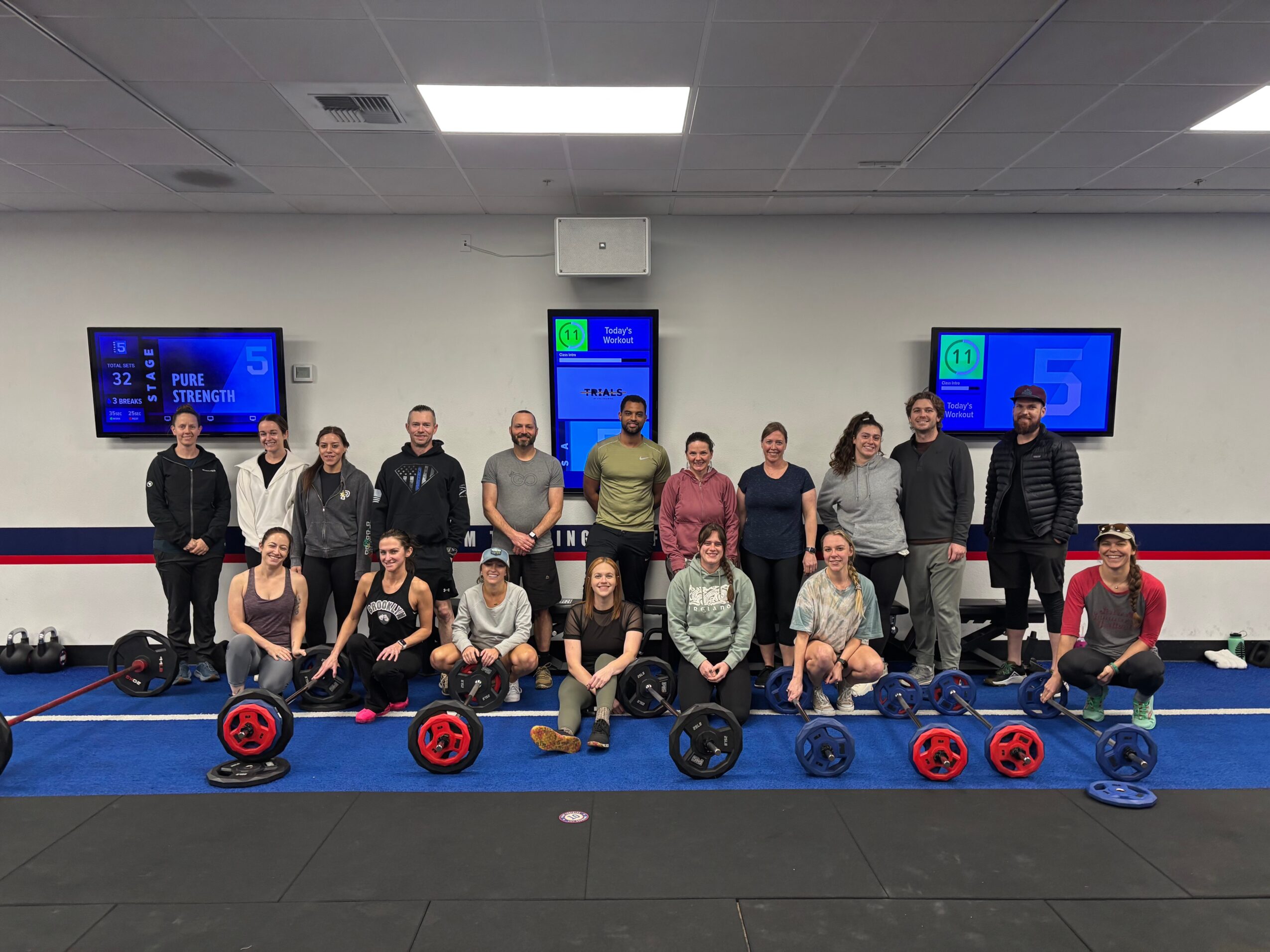 Group of people in a gym with weightlifting equipment, two screens on the wall, and a blue mat on the floor.