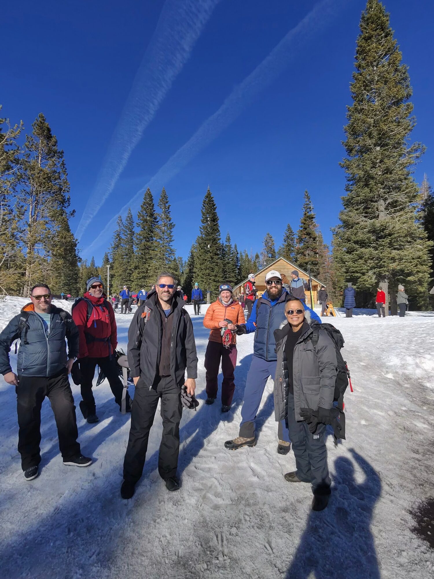 Group of people standing outdoors in snowy landscape with tall trees and blue sky, some wearing sunglasses and backpacks.
