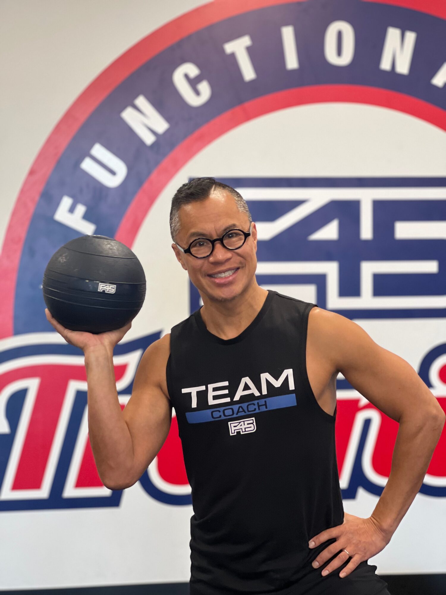 Person smiling, holding a medicine ball, standing in front of a fitness-themed background with text and logos.