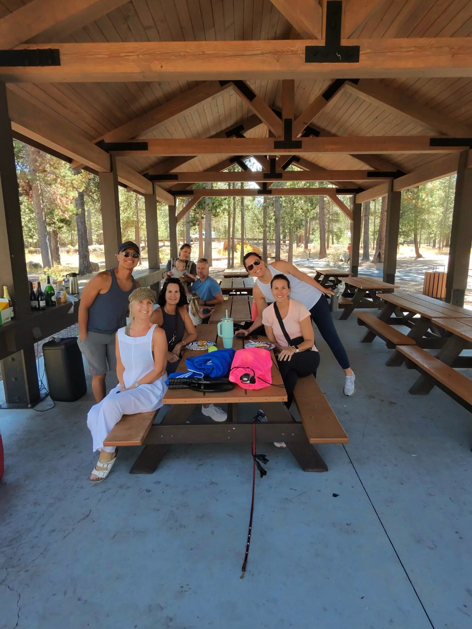Group of seven people sitting at picnic tables in a pavilion outdoors, with trees in the background.