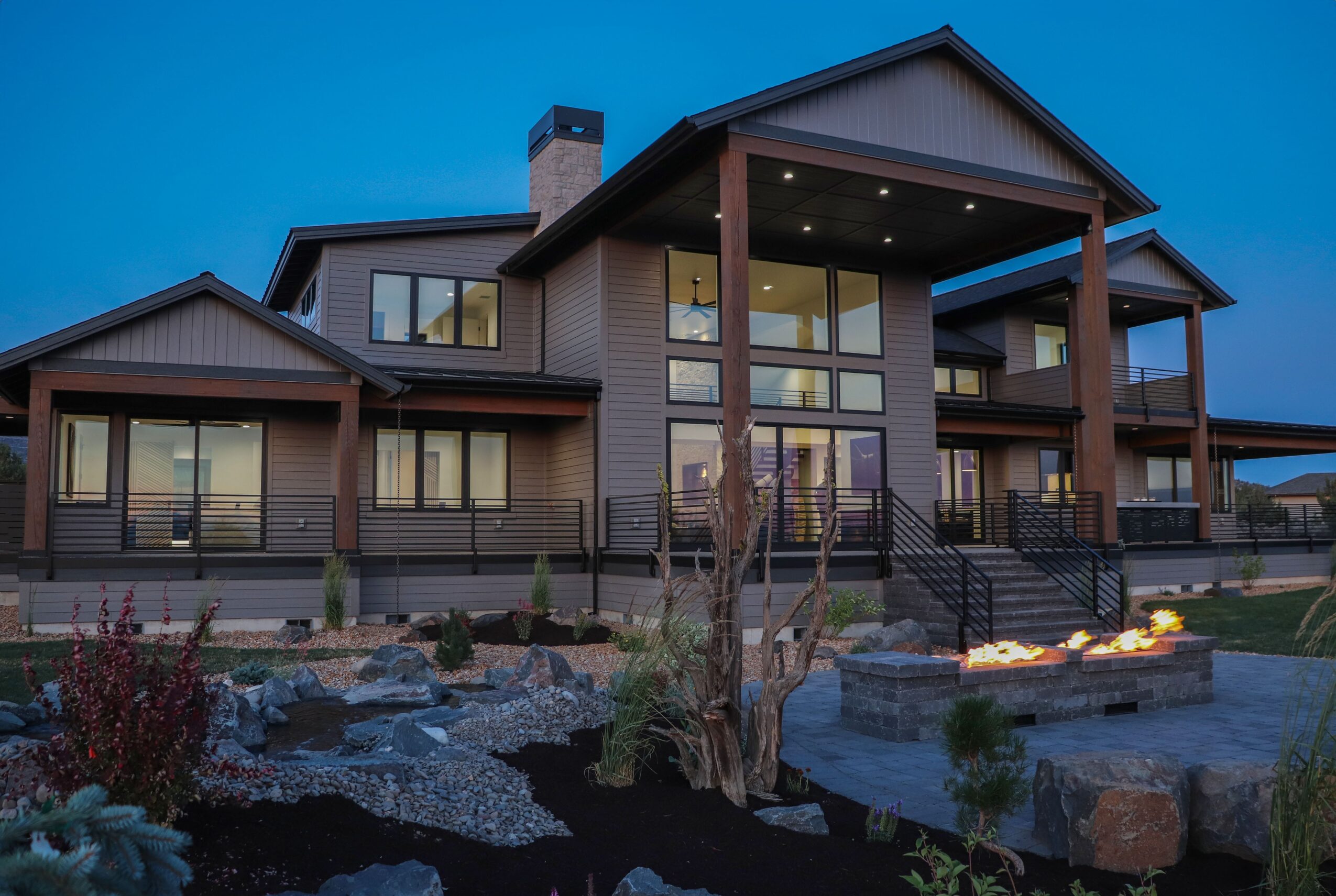 Large modern house with multiple levels, balconies, and large windows, illuminated at dusk with landscaped yard and pathway.