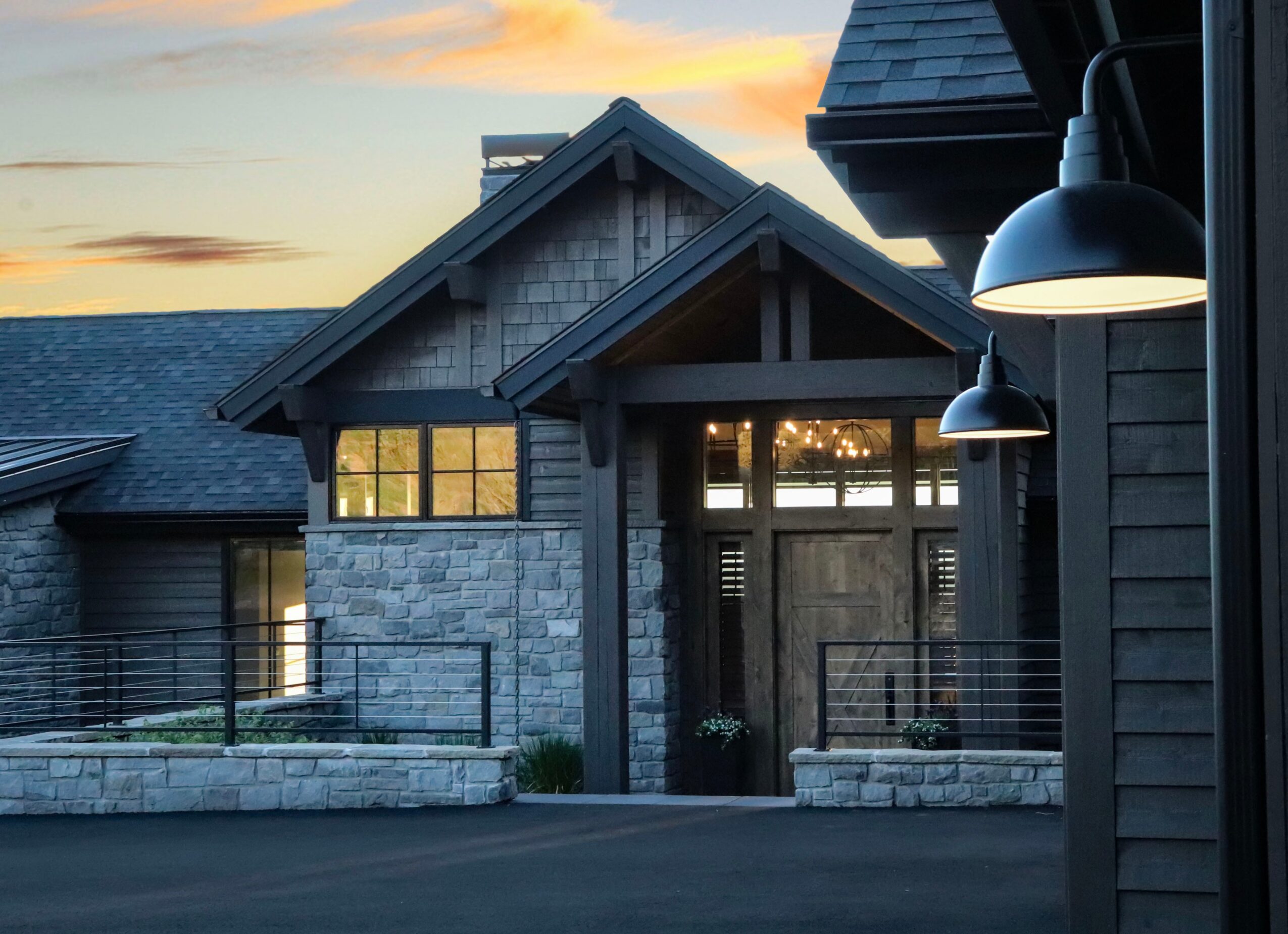 Exterior of a modern house with stone and wood siding, illuminated lamps, and a sunset sky in the background.