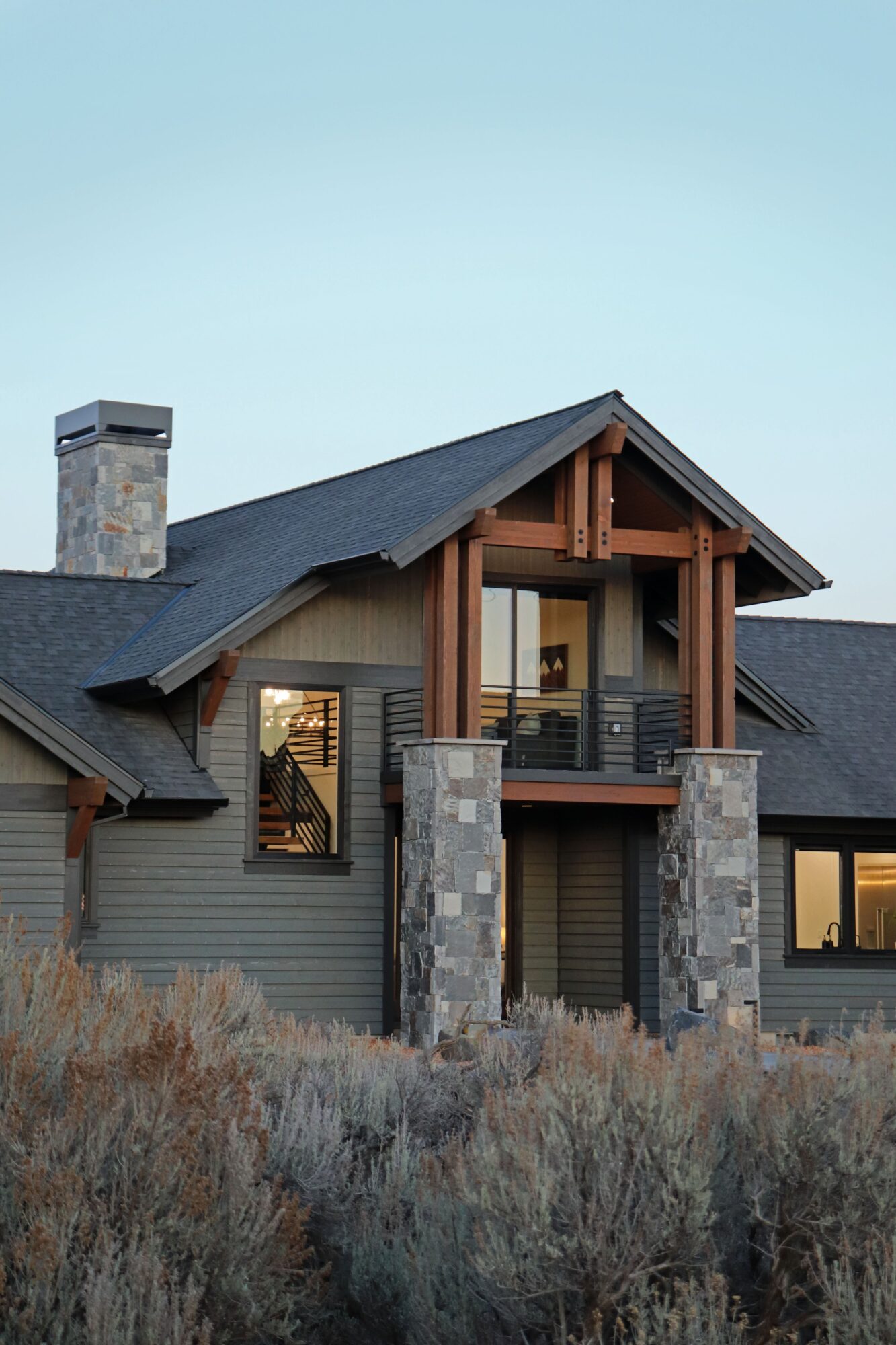 Modern house with stone pillars, wooden beams, and large windows, set against a clear sky and natural landscape.