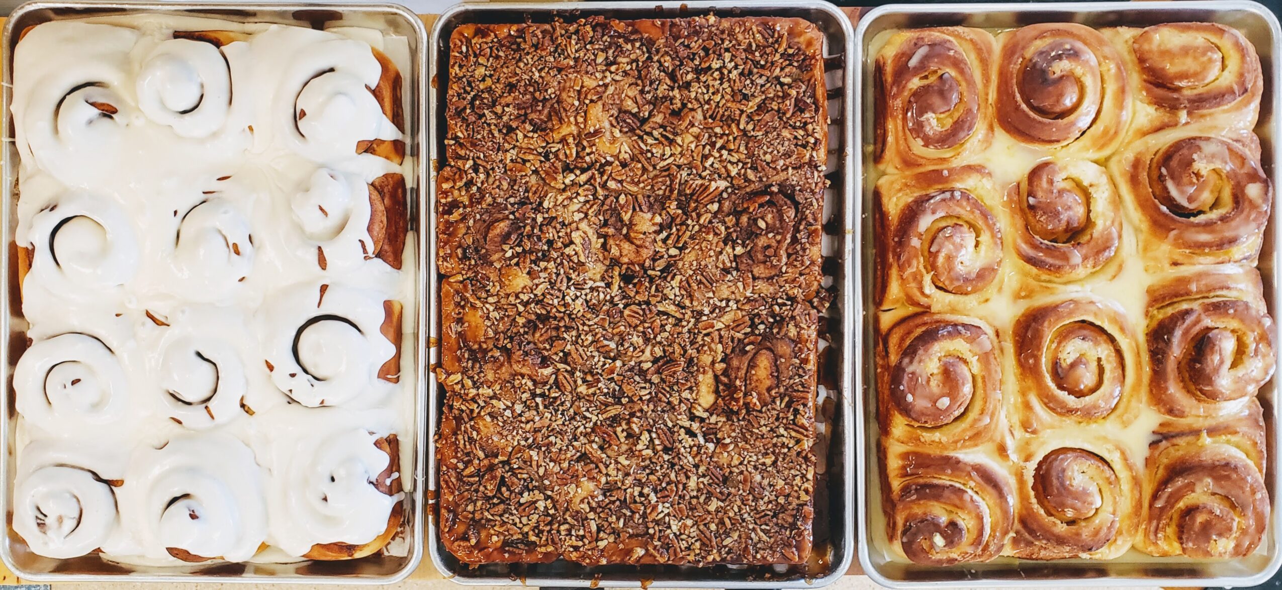 Three trays of baked goods: a tray of cinnamon rolls with icing, a tray of crumb-topped dessert, and a tray of cinnamon rolls with glaze.