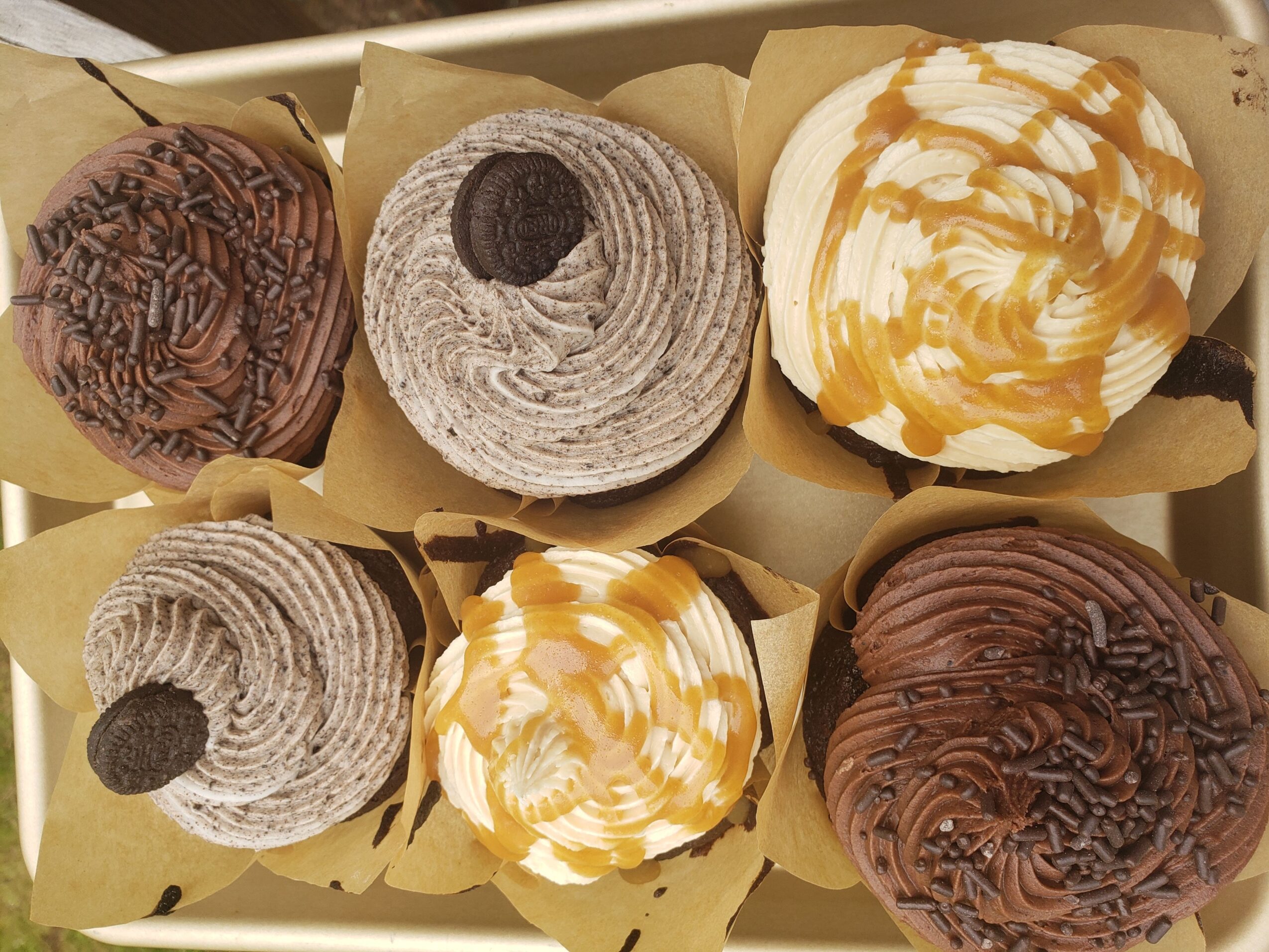 Six cupcakes with various frosting designs and toppings arranged in two rows, on yellow paper liners.