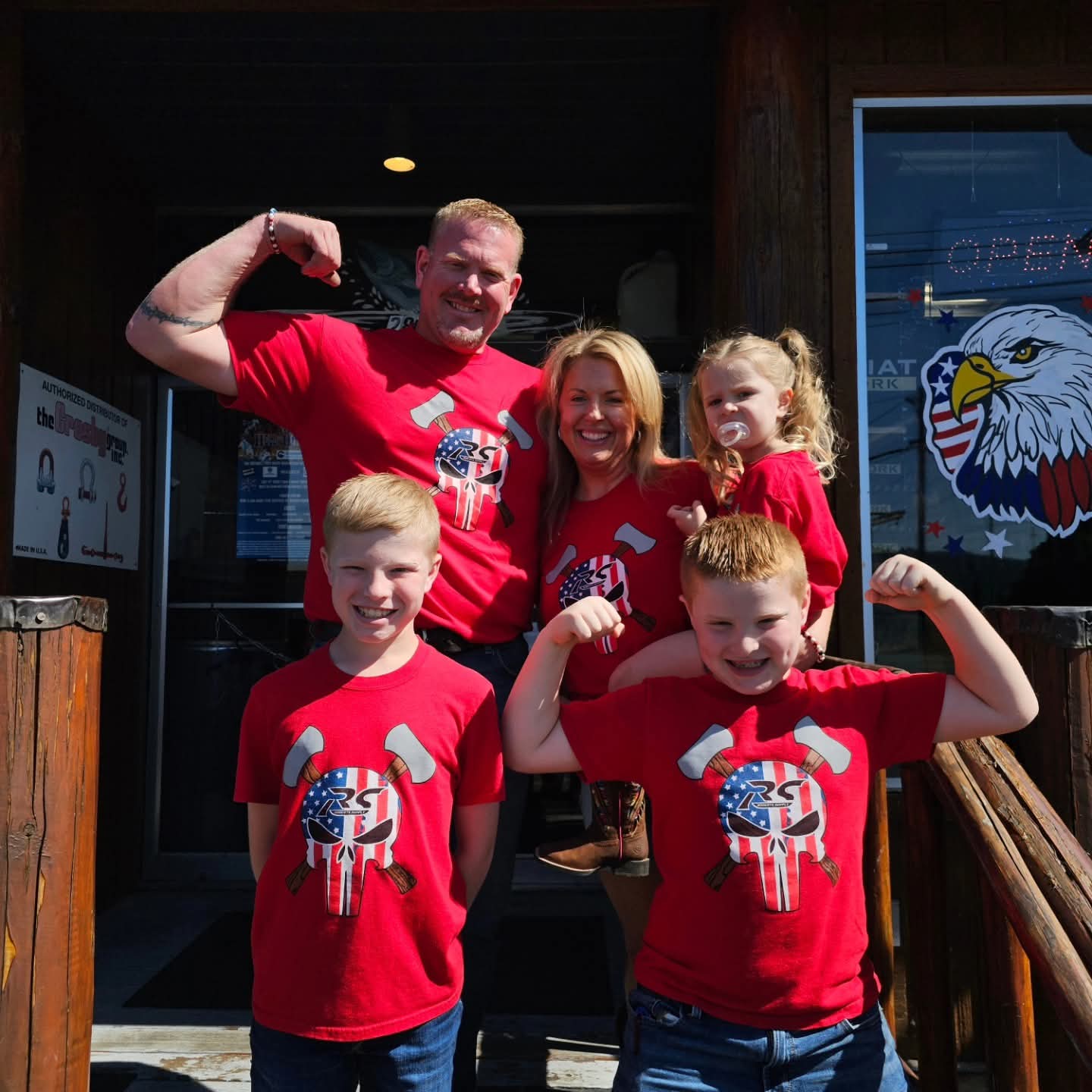 Group of five people, two adults and three children, smiling and flexing muscles outside a building, all wearing red shirts with a dog graphic.