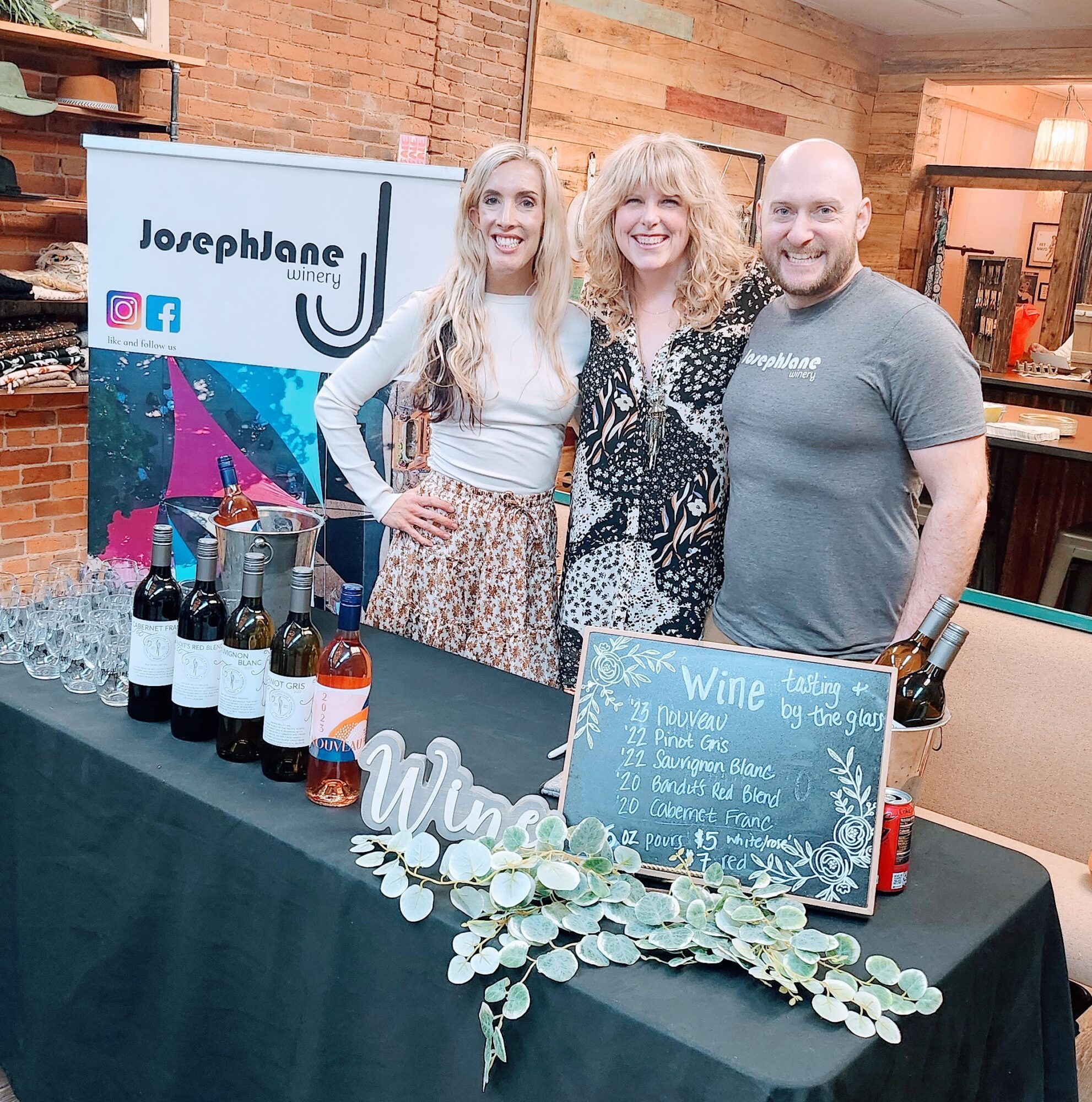 Three people standing behind a table with wine bottles, a chalkboard sign, and decorative greenery in a cozy indoor setting.
