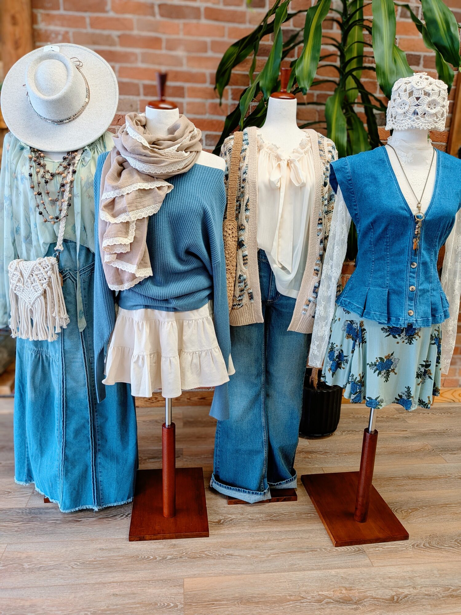 Four mannequins dressed in blue and white summer outfits, with hats and accessories, against a brick wall and plant background.
