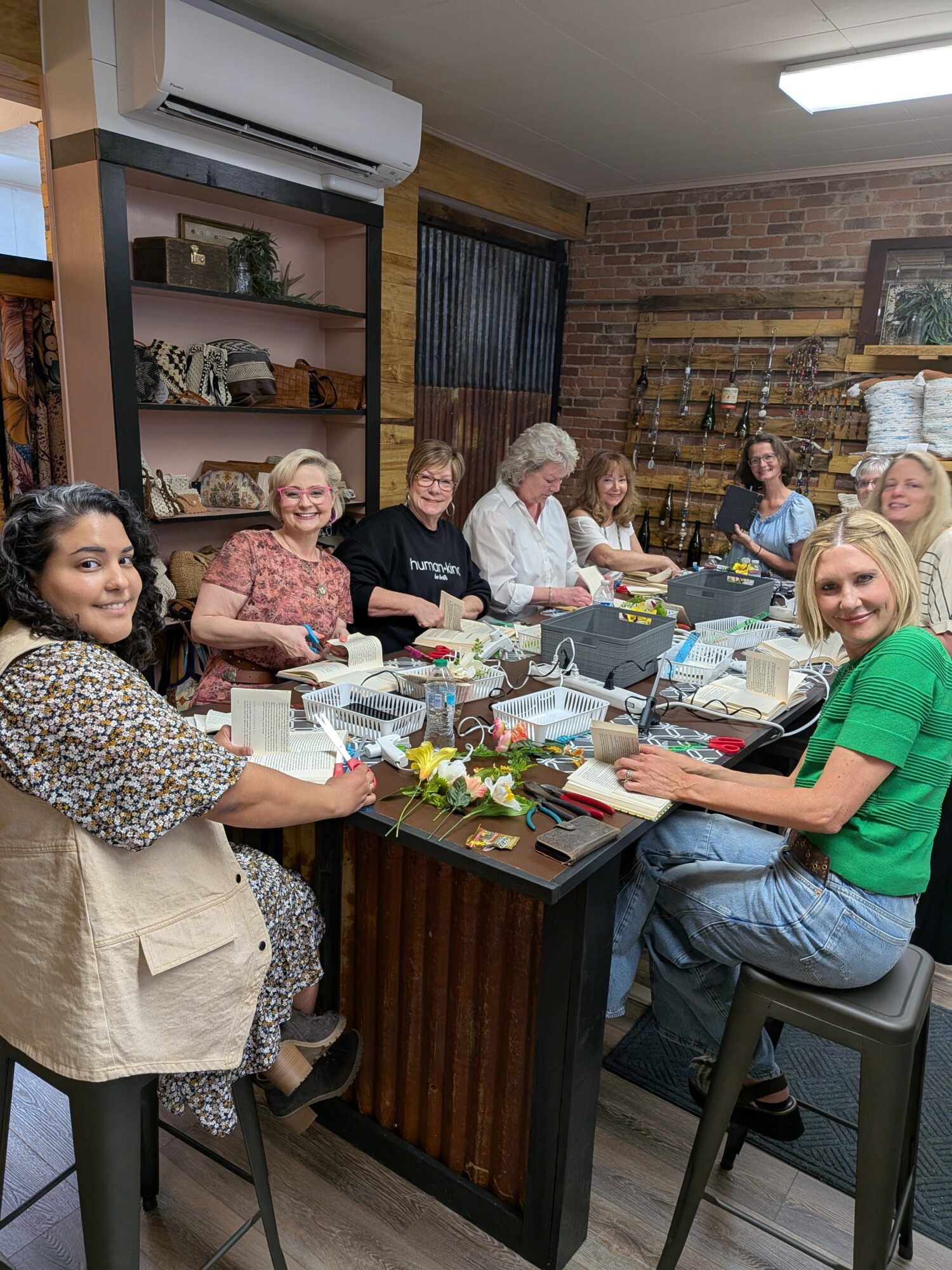 Group of women sitting around a table with flowers, papers, and office supplies in a room with brick wall and shelves.