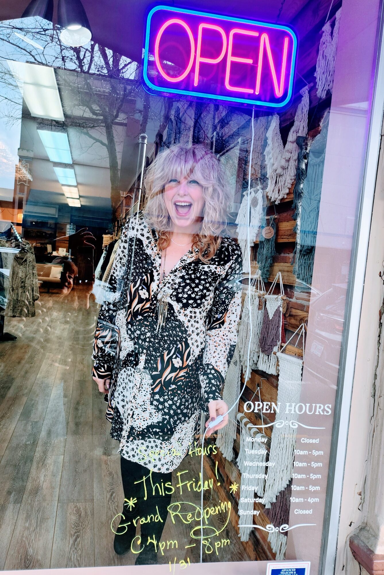 Woman with curly hair smiling behind a glass window with an 'OPEN' sign, decorated with hanging ornaments, inside a store.