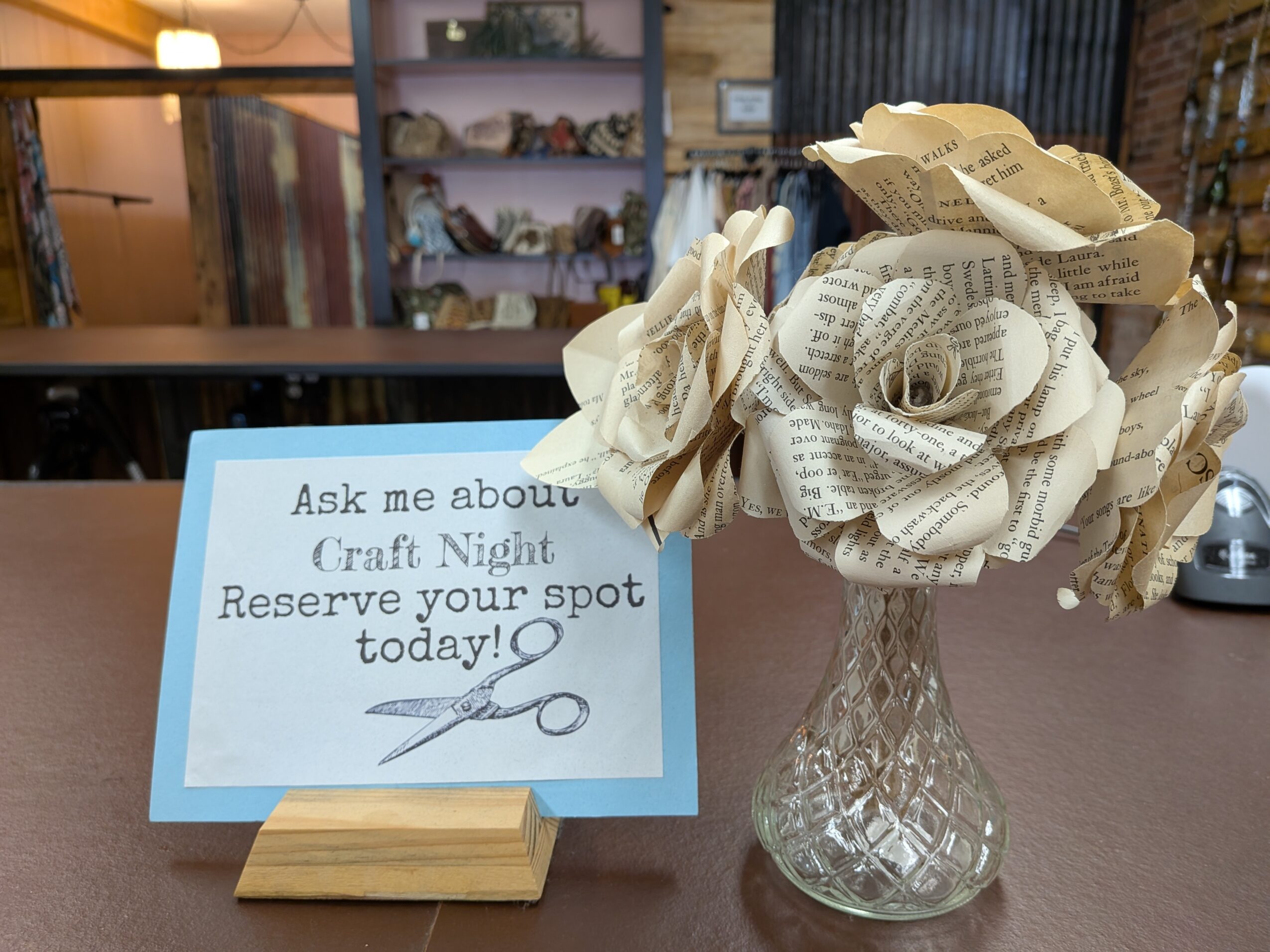 Paper flower made from rolled newspaper in a glass vase on a table, with a sign inviting questions about craft night.
