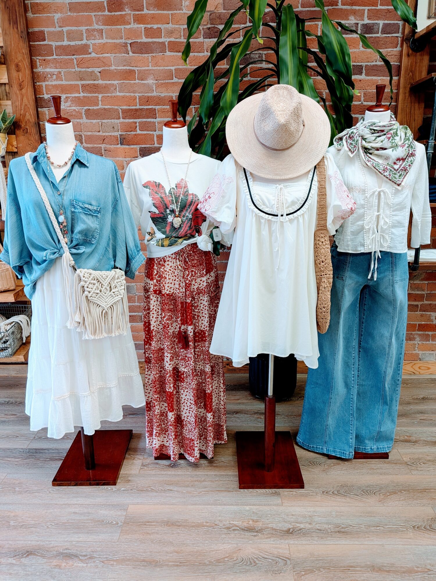 Four mannequins display casual summer outfits with dresses, tops, pants, and accessories against a brick wall and green plant.