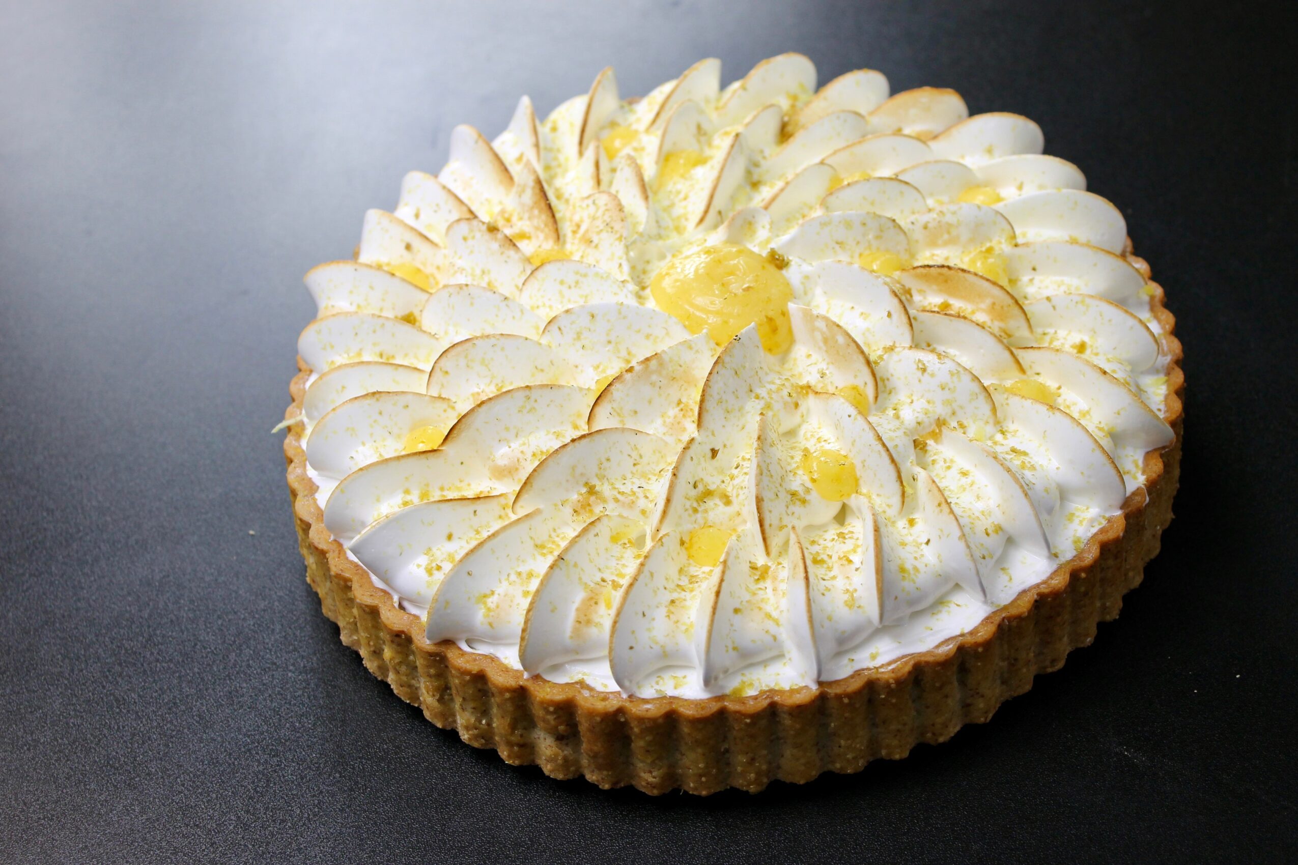 Round tart with white cream and sliced fruit on top, on a dark surface.