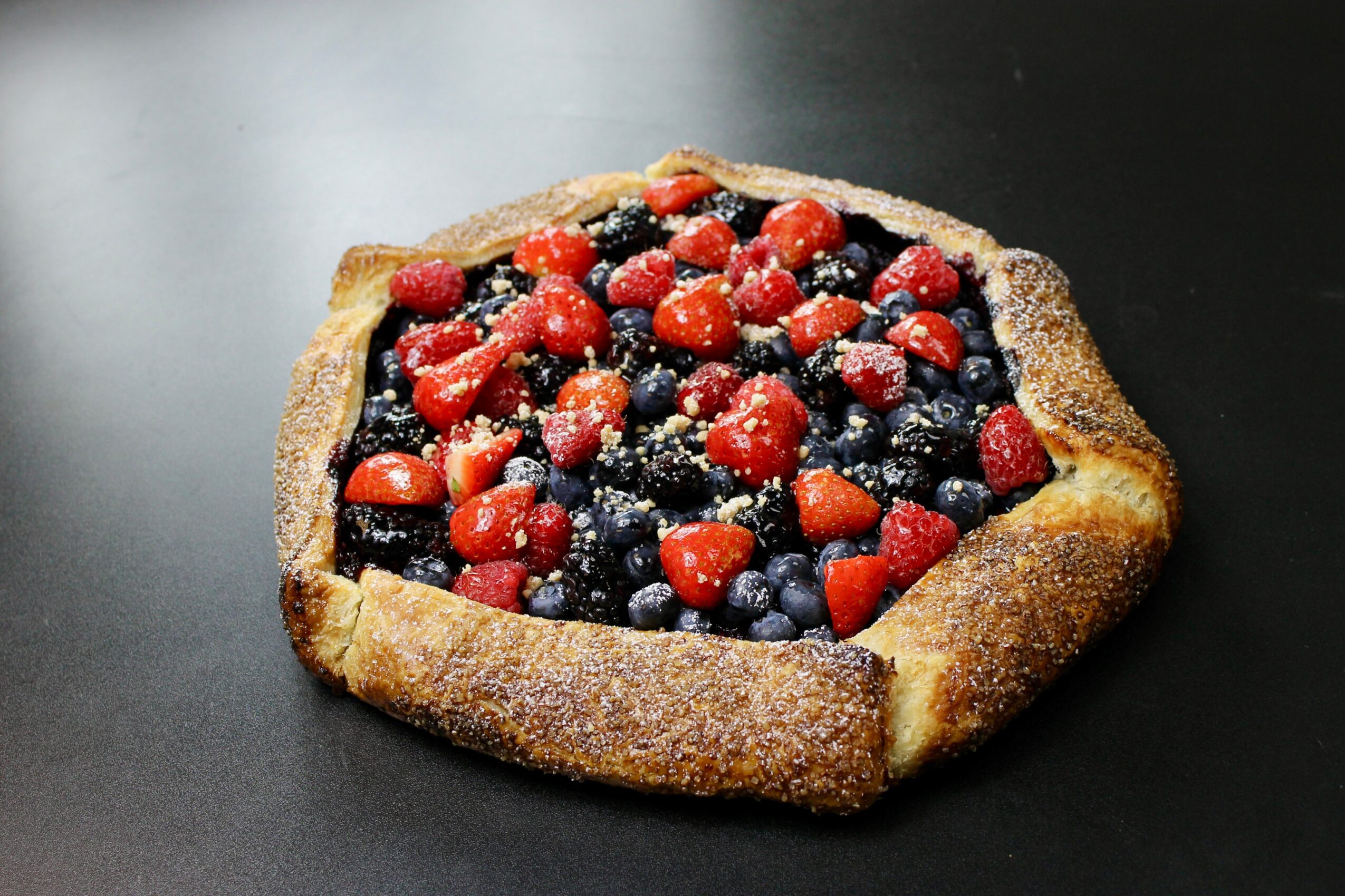 Square fruit tart with mixed berries on a dark surface, crust edges slightly raised, berries appear fresh and colorful.
