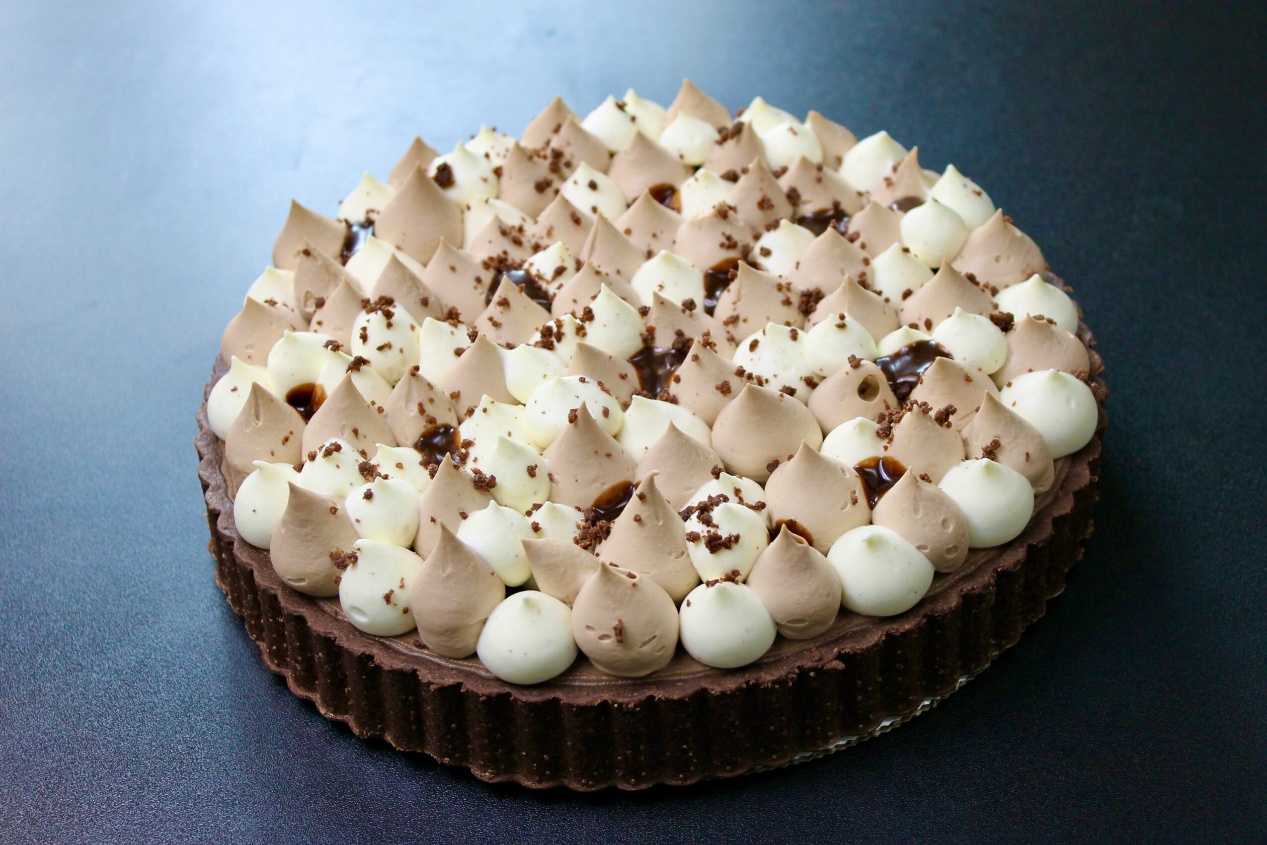 Round chocolate cake topped with white and light brown cream swirls and chocolate shavings, on a dark surface.