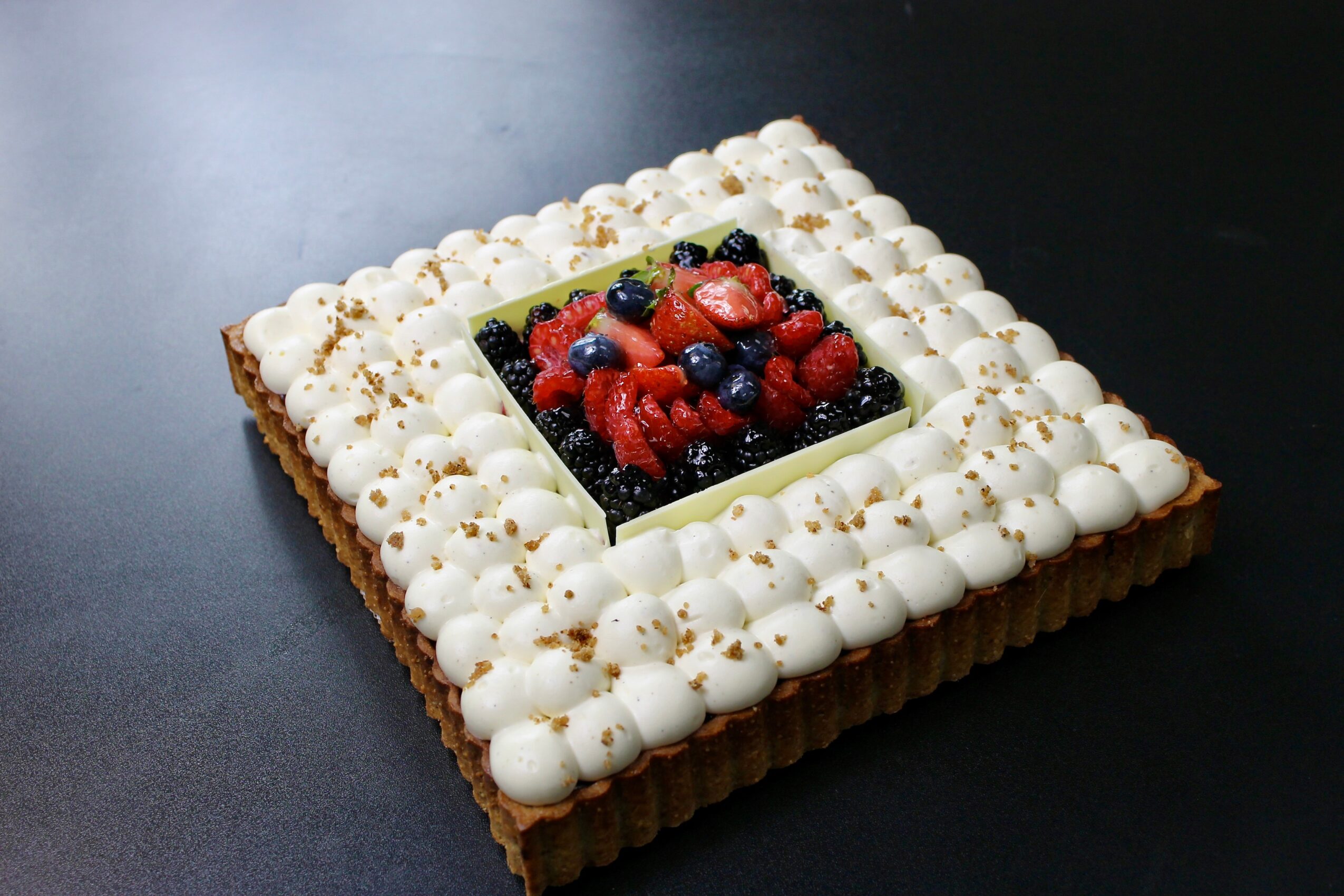 Square fruit cake with white cream border and mixed berries in a central square opening, on a dark surface.
