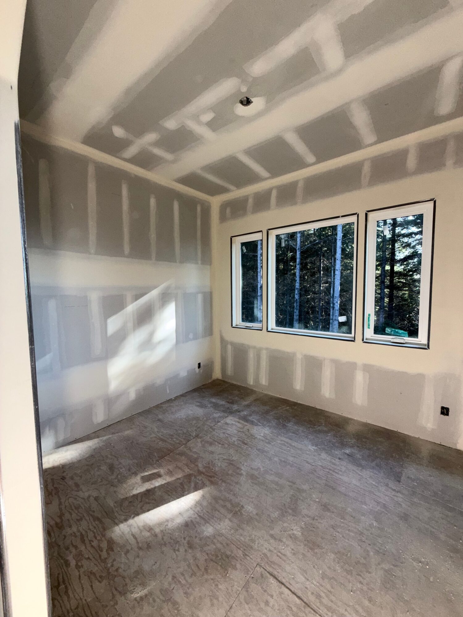 Empty room with three large windows, drywall with visible seams, and a carpeted floor with a vent, sunlight streaming in.