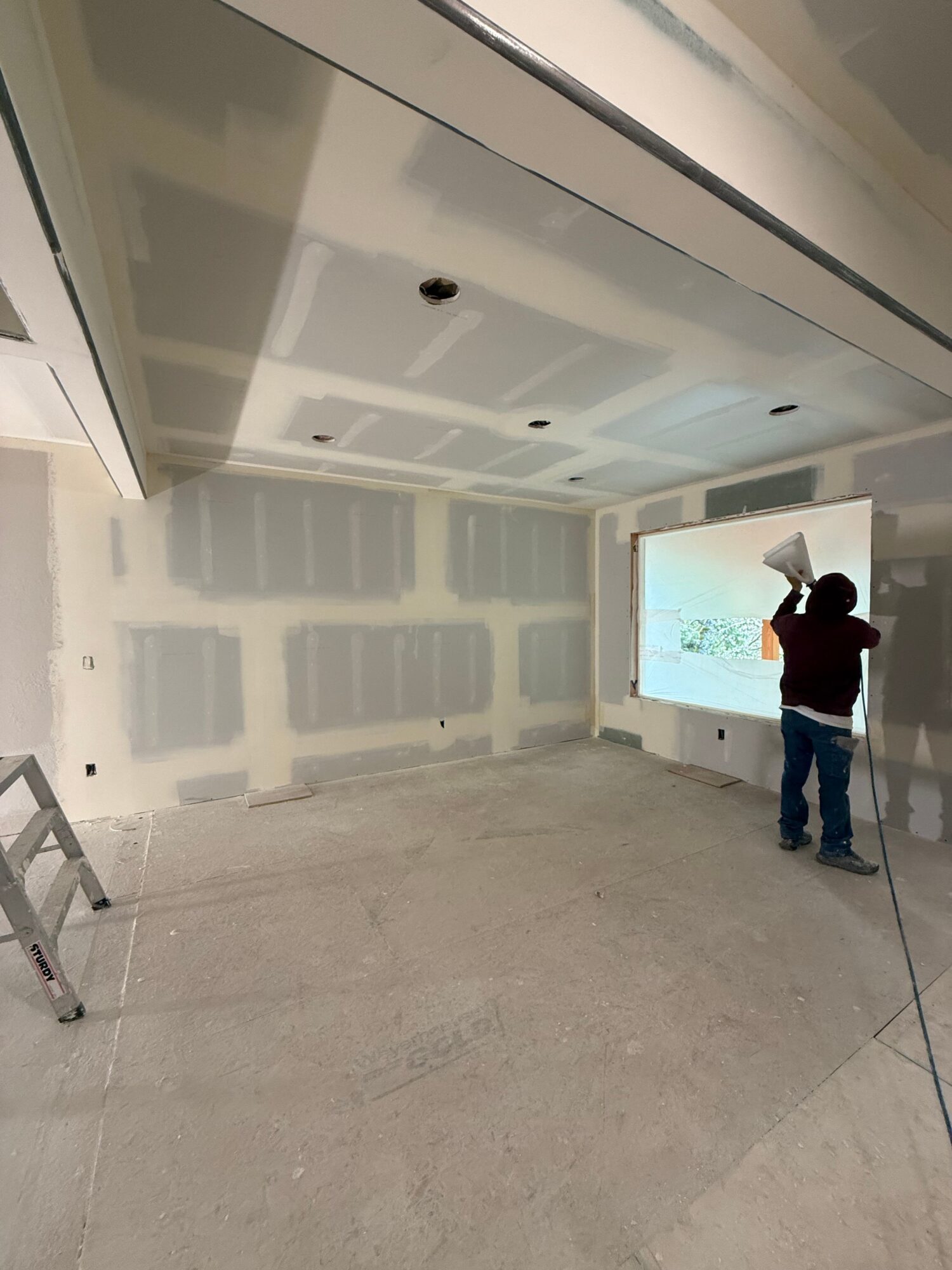 Person standing in a room with unfinished walls, looking at a screen on the wall, with a ladder nearby.