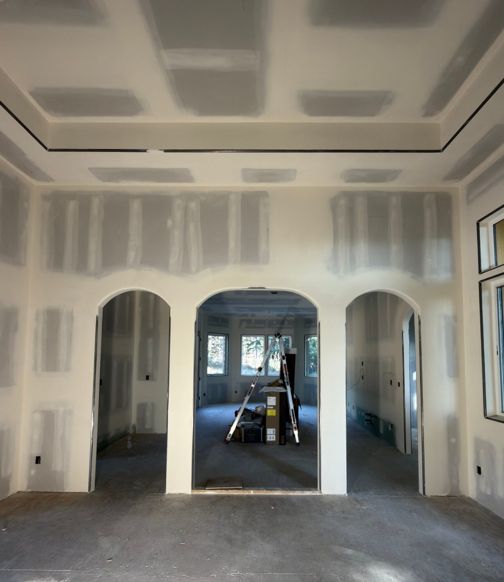 Room with three arches, drywall installation in progress, window at the back, construction materials in the center, ceiling partially finished.
