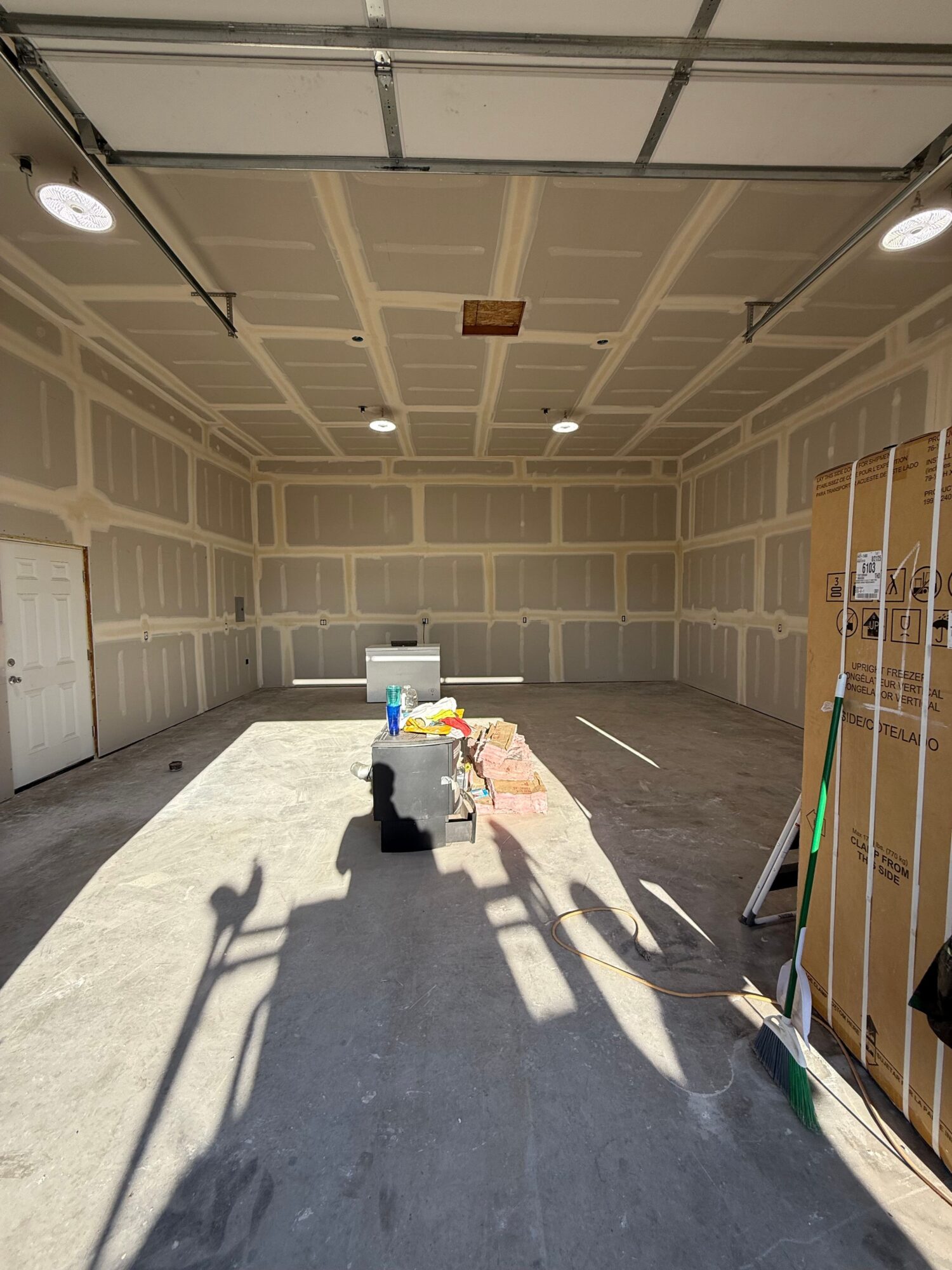 Garage interior with unfinished walls, ceiling, and a concrete floor. Sunlight casts shadows on the floor. Items are near the back wall.