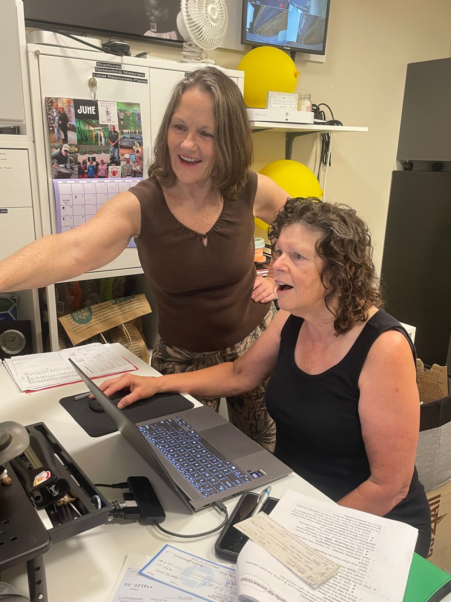 Two women working together at a desk with a laptop, papers, and office supplies, smiling and engaged in conversation.
