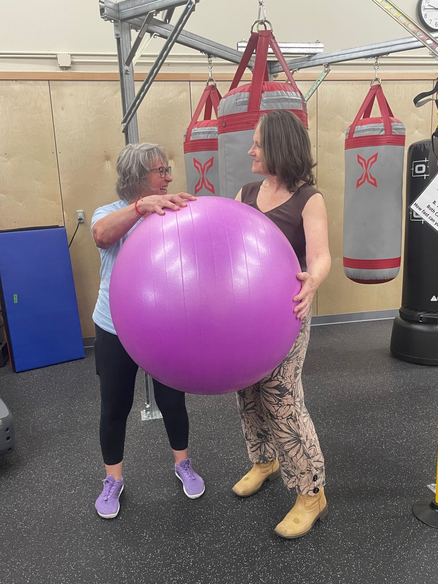 Two women are exercising with a large purple ball in a gym, one woman is holding the ball while the other supports it.