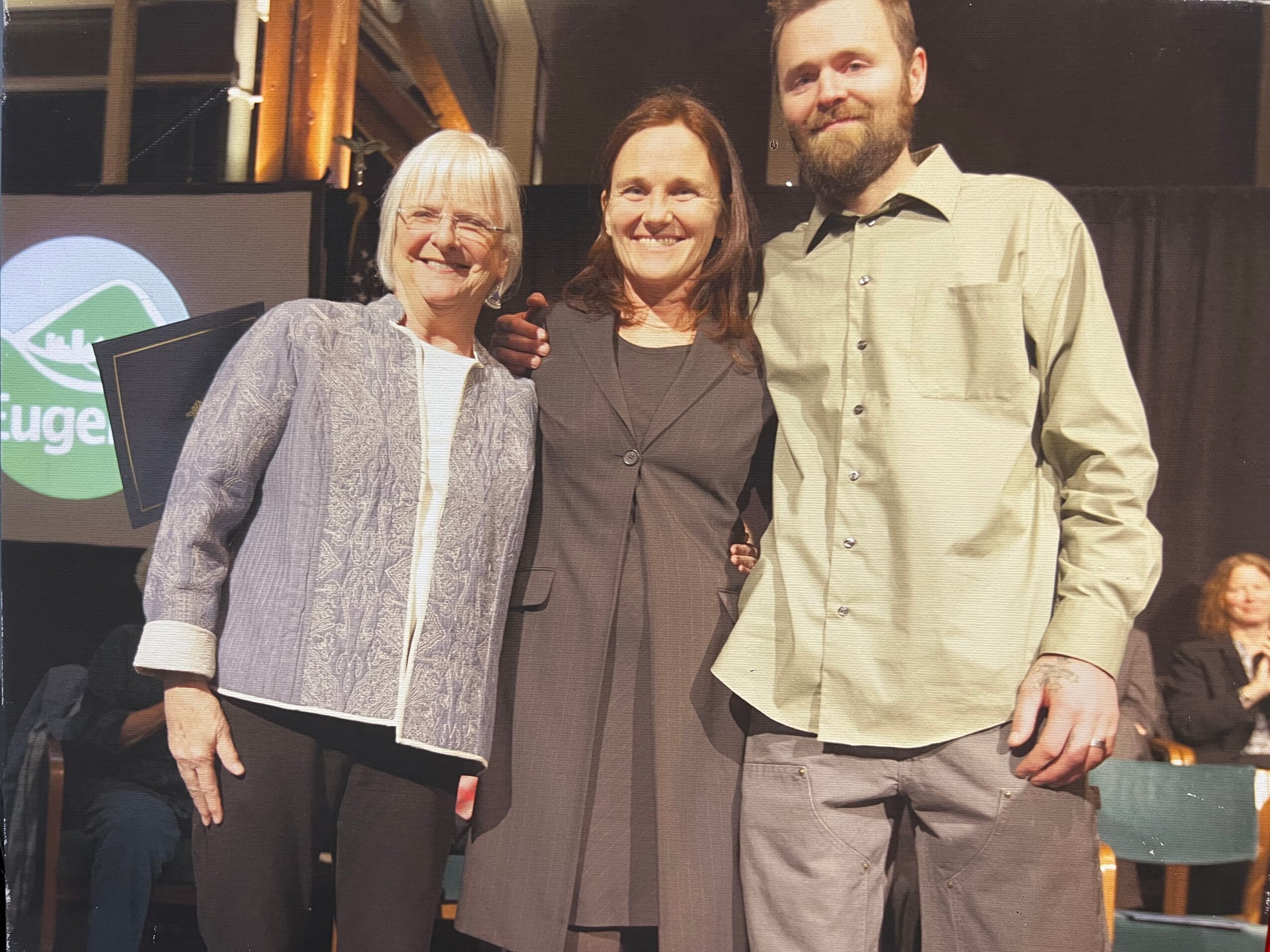 Three people standing together, smiling, in an indoor setting with a stage and audience in the background.