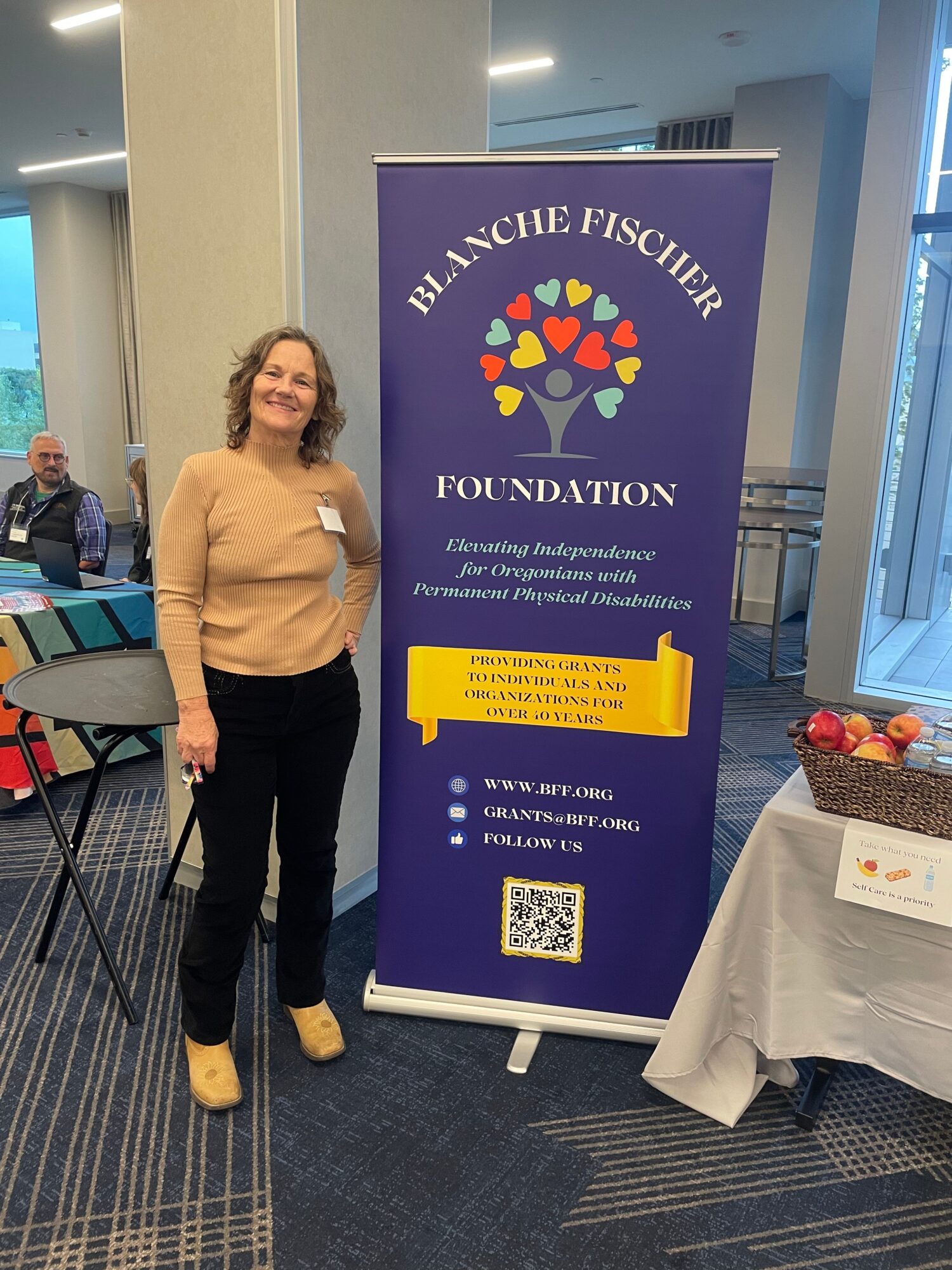 Woman standing next to a purple and yellow sign at an indoor event, with a table and a QR code visible.